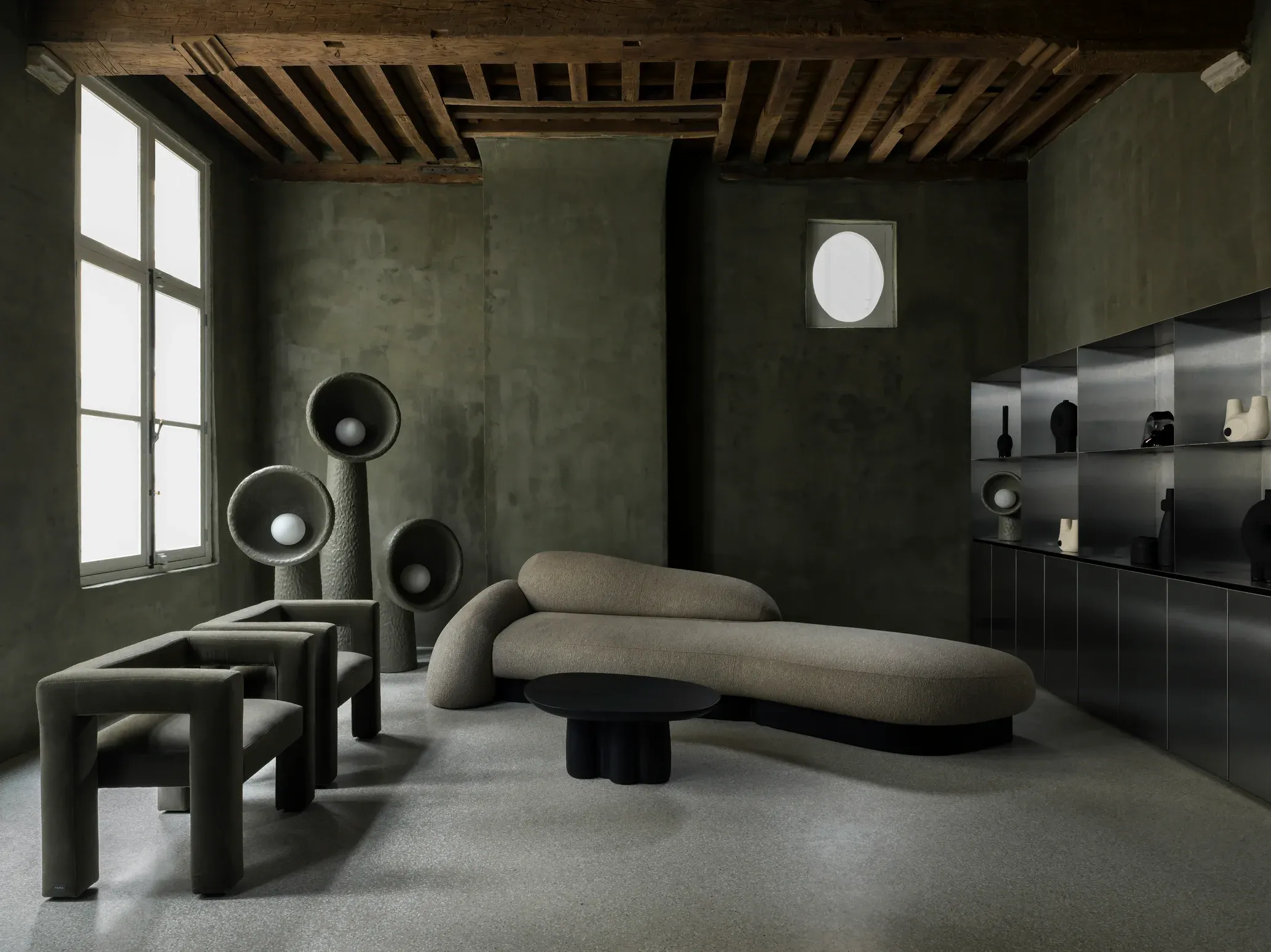 Faina, Europe — Sculptural furniture in natural linen and black materials arranged in brutalist interior with concrete walls and wooden ceiling beams.