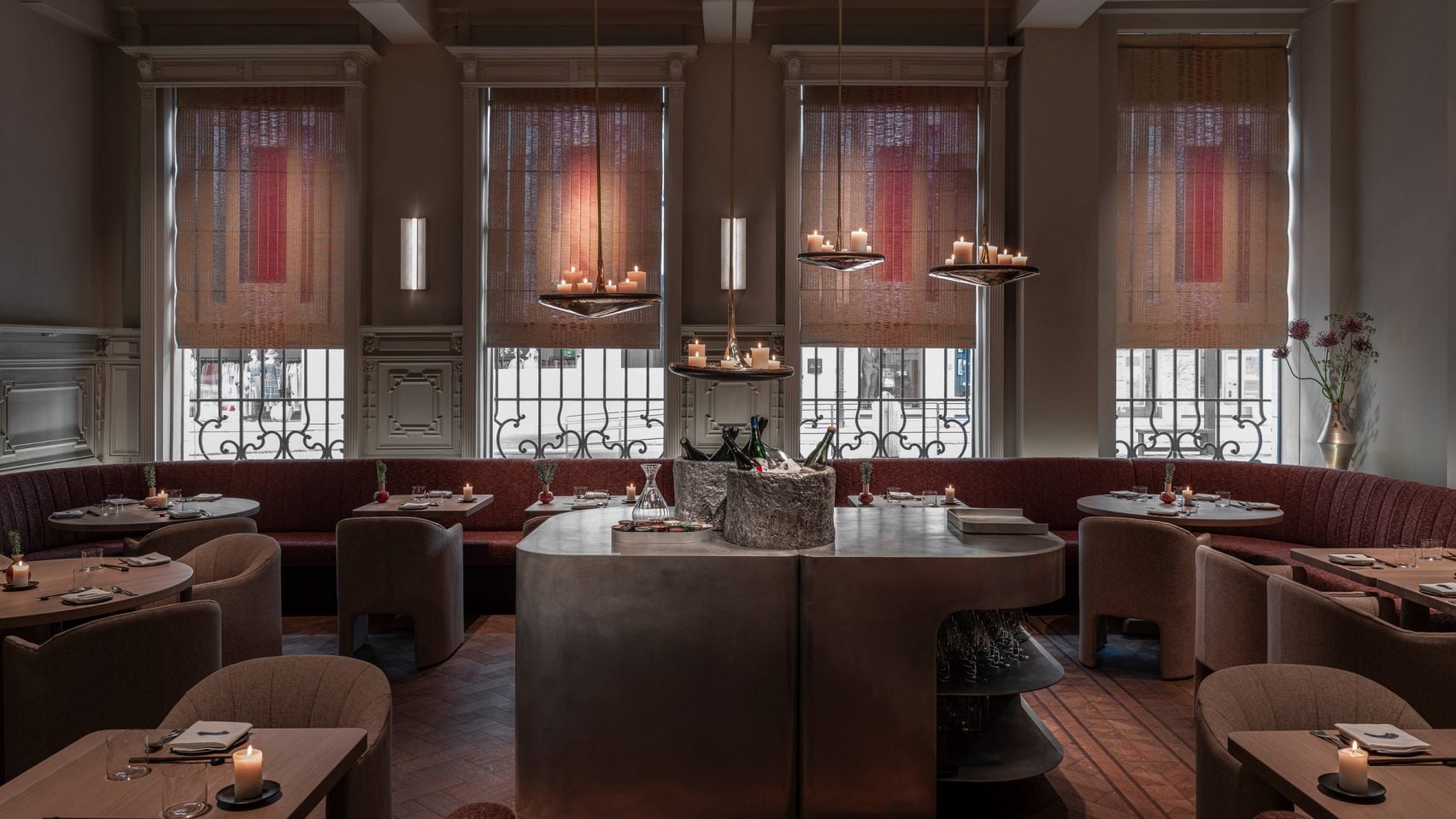 Blueness, Europe — Candlelit dining room with burgundy banquettes, sculptural bronze tables, floor-to-ceiling windows overlooking Parisian balconies, warm ambient lighting.