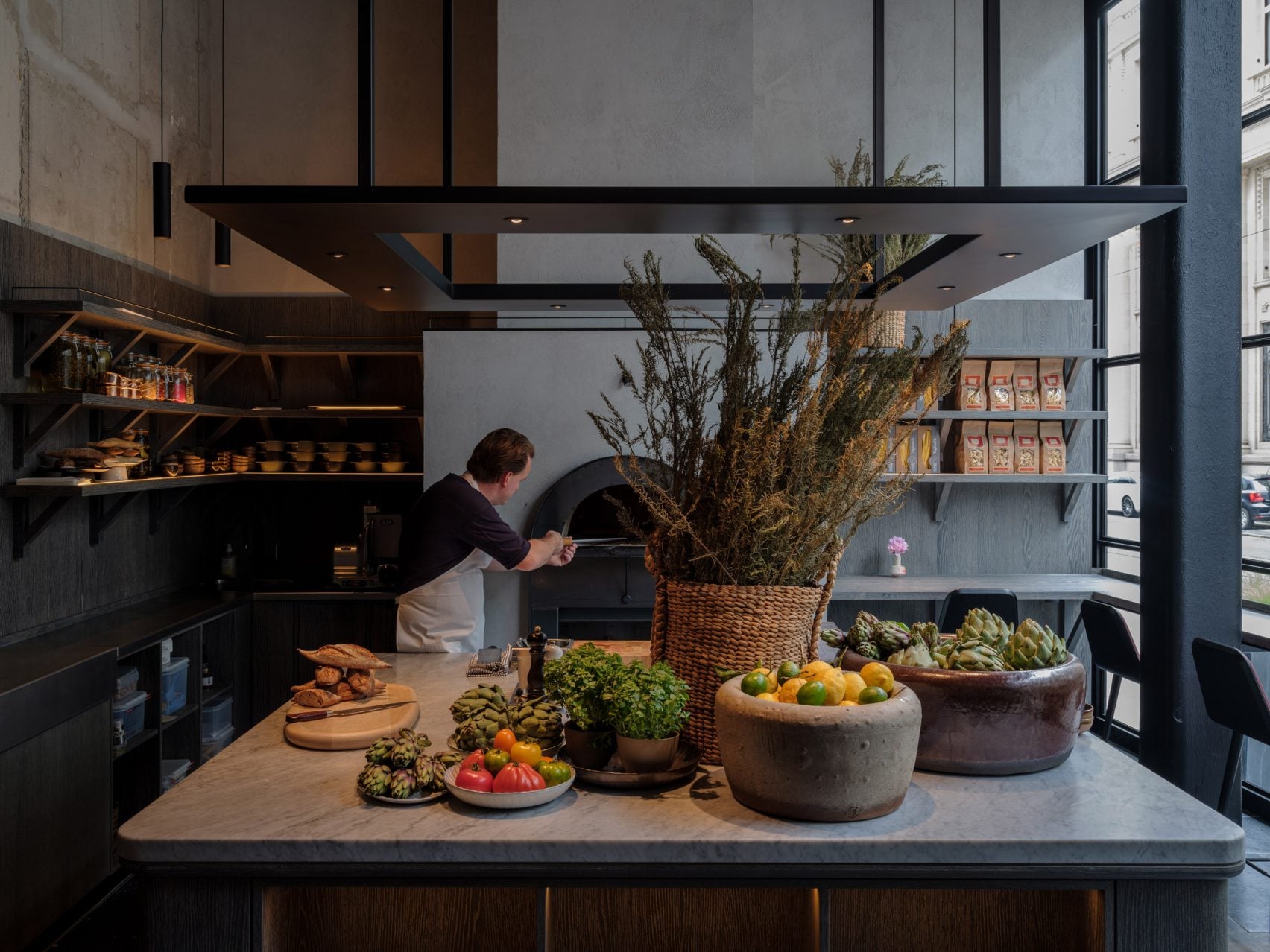 Le Pristine, Europe — Modern kitchen with chef at open hearth, dried grasses in woven basket, fresh produce in concrete bowls, black steel beams, natural light from floor-to-ceiling windows.