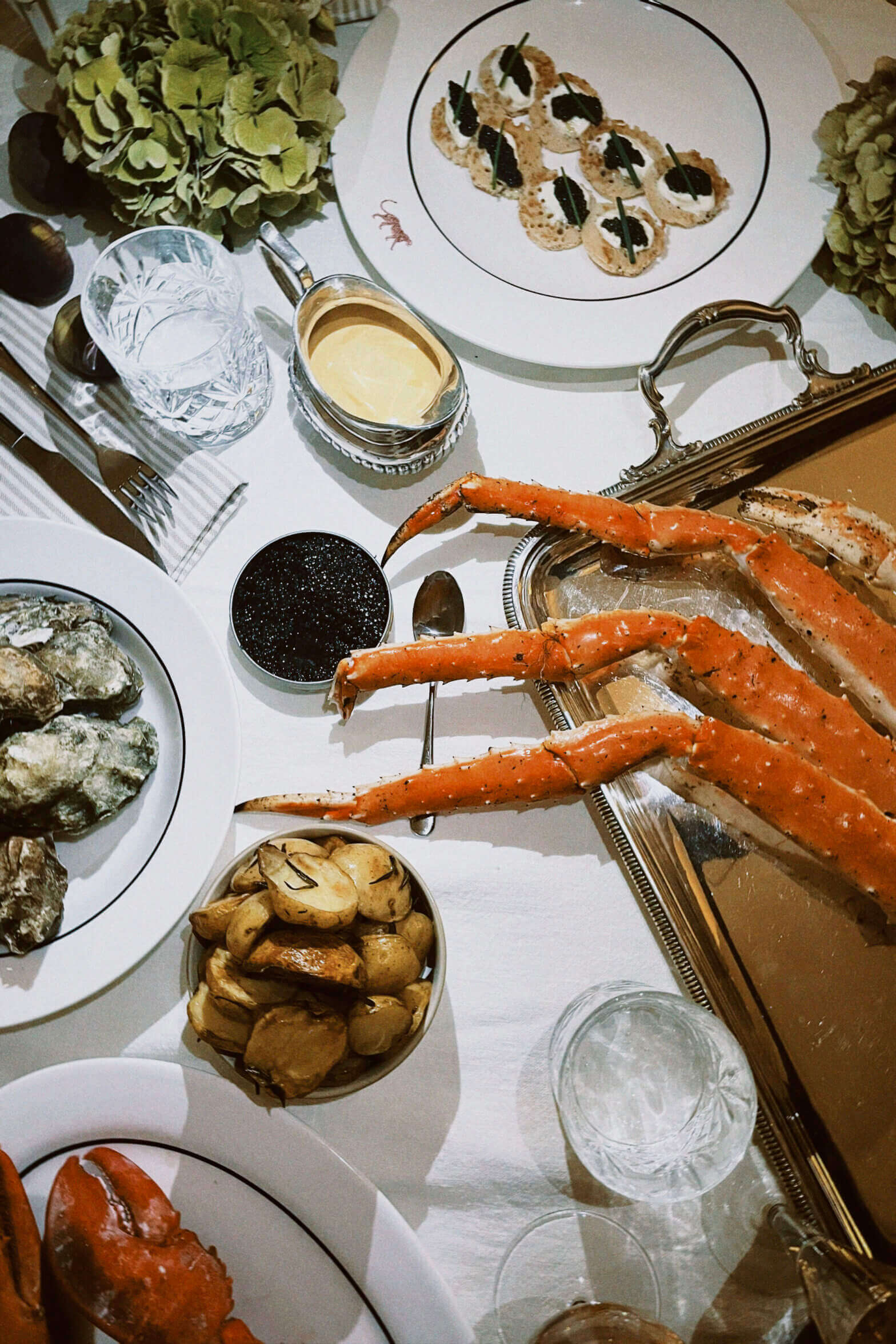 Palm Heights, North America — Overhead flatlay of luxury seafood service with king crab legs, oysters, caviar, canapés, and champagne on white tablecloth.