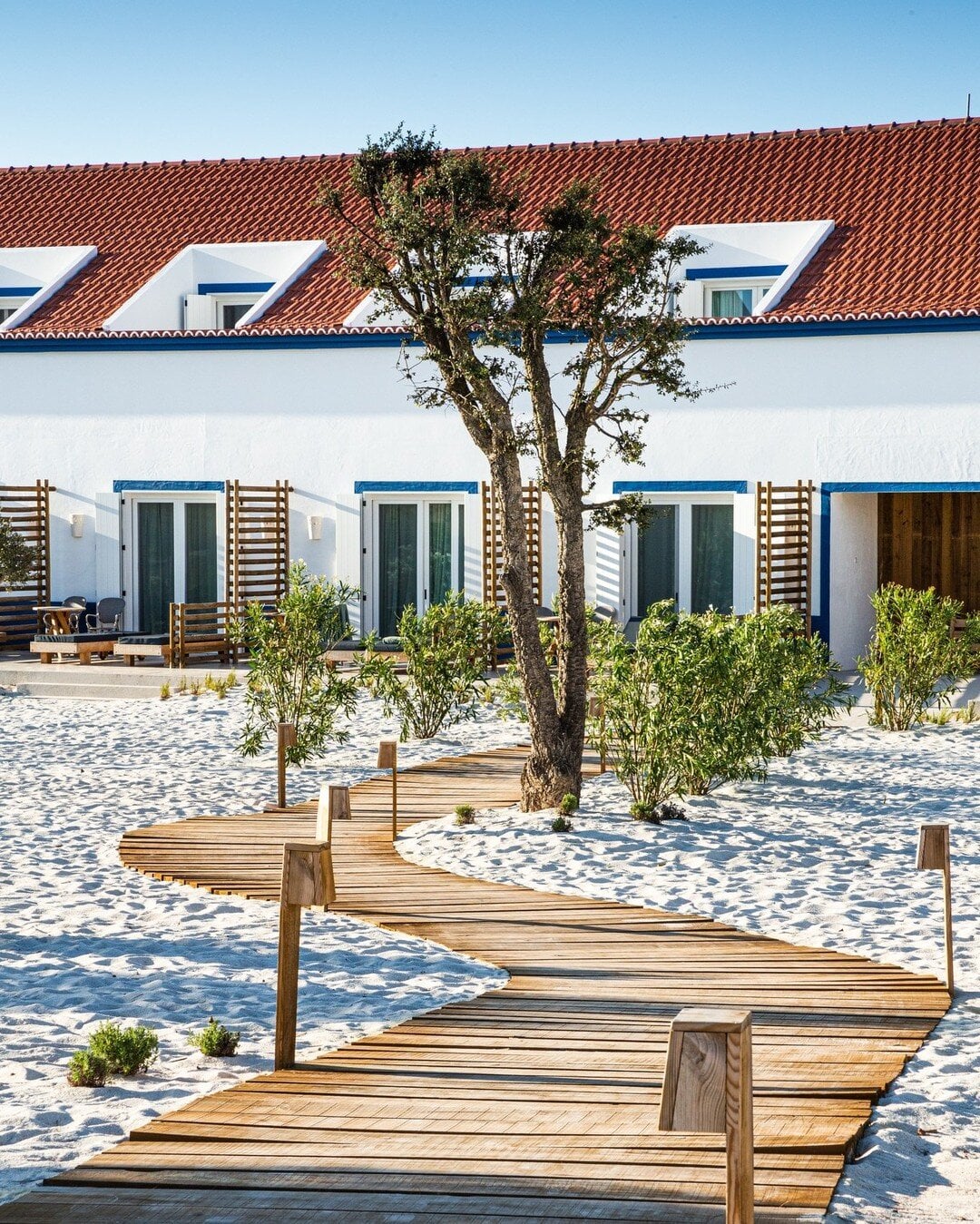 Quinta da Comporta, Portugal — Wooden boardwalk crossing white sand beach toward whitewashed cottages with terracotta roofs and blue accents, gnarled tree in foreground.