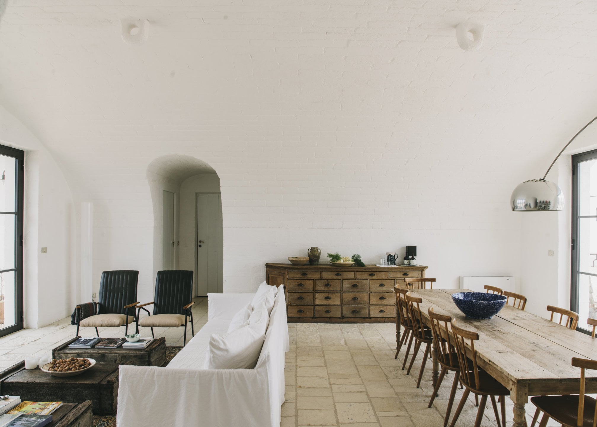 Masseria Moroseta, Europe — Whitewashed stone interior with arched doorway, wooden dining table, linen sofa, arc lamp, and minimal Mediterranean furnishings.