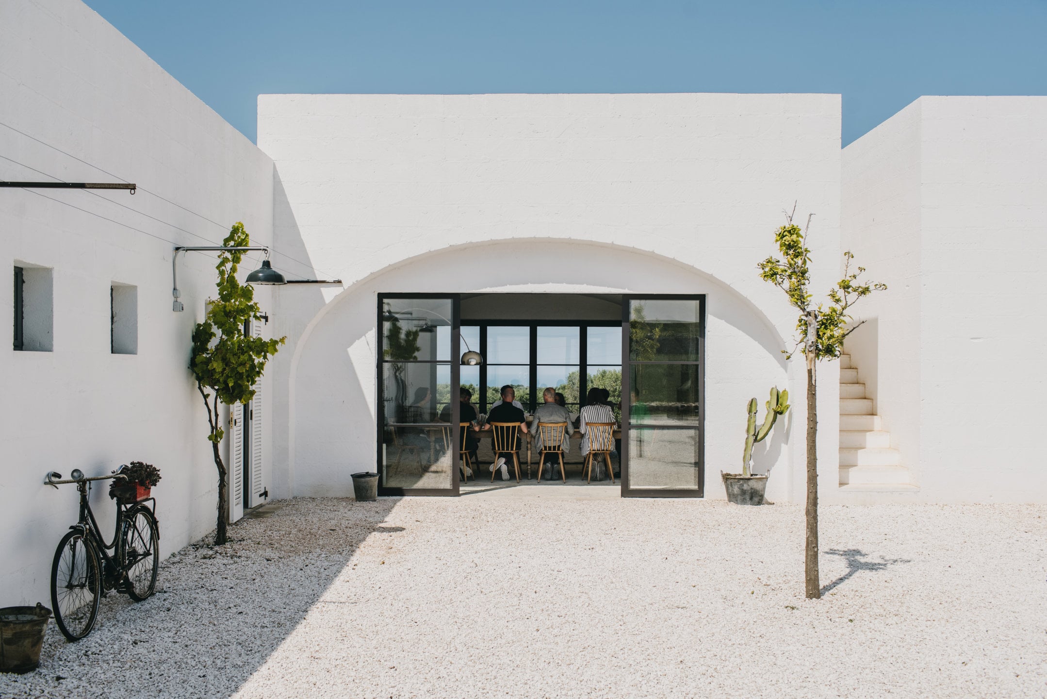 Masseria Moroseta, Europe — Whitewashed courtyard with vaulted archway, black-framed glass doors, gravel terrace, minimal potted plants, and diners visible inside.