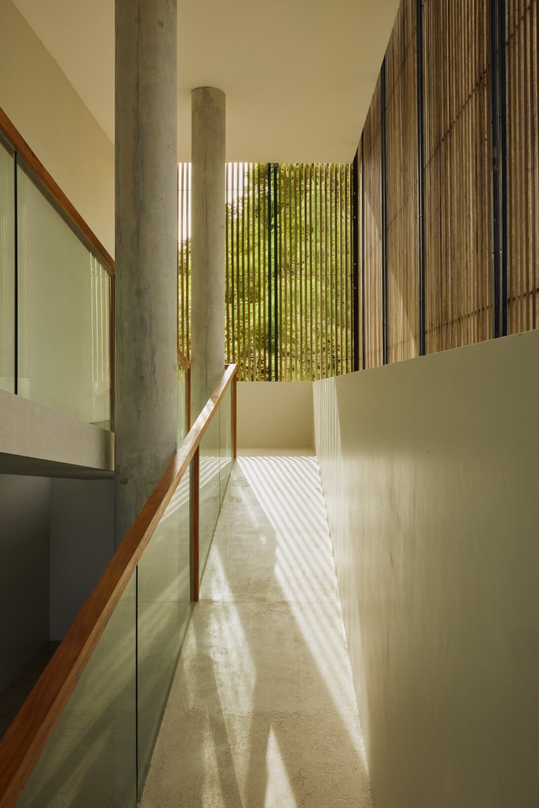 Innit Lombok, Asia — Concrete columns and wooden handrail frame minimalist corridor with vertical wood slats filtering warm sunlight across pale floors.