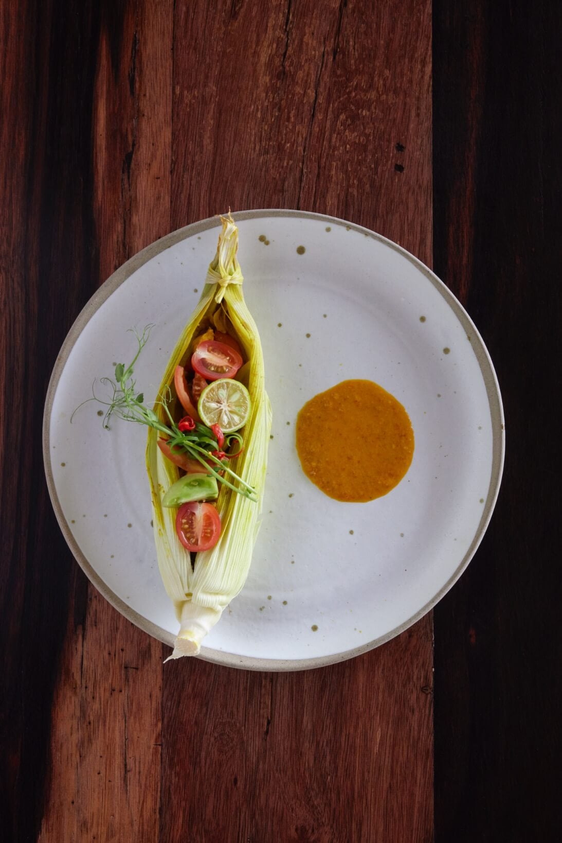 Innit Lombok, Indonesia — Steamed fish wrapped in corn husks with tomato, lime, and herbs, served with turmeric sauce on speckled ceramic plate, overhead view on wood table.
