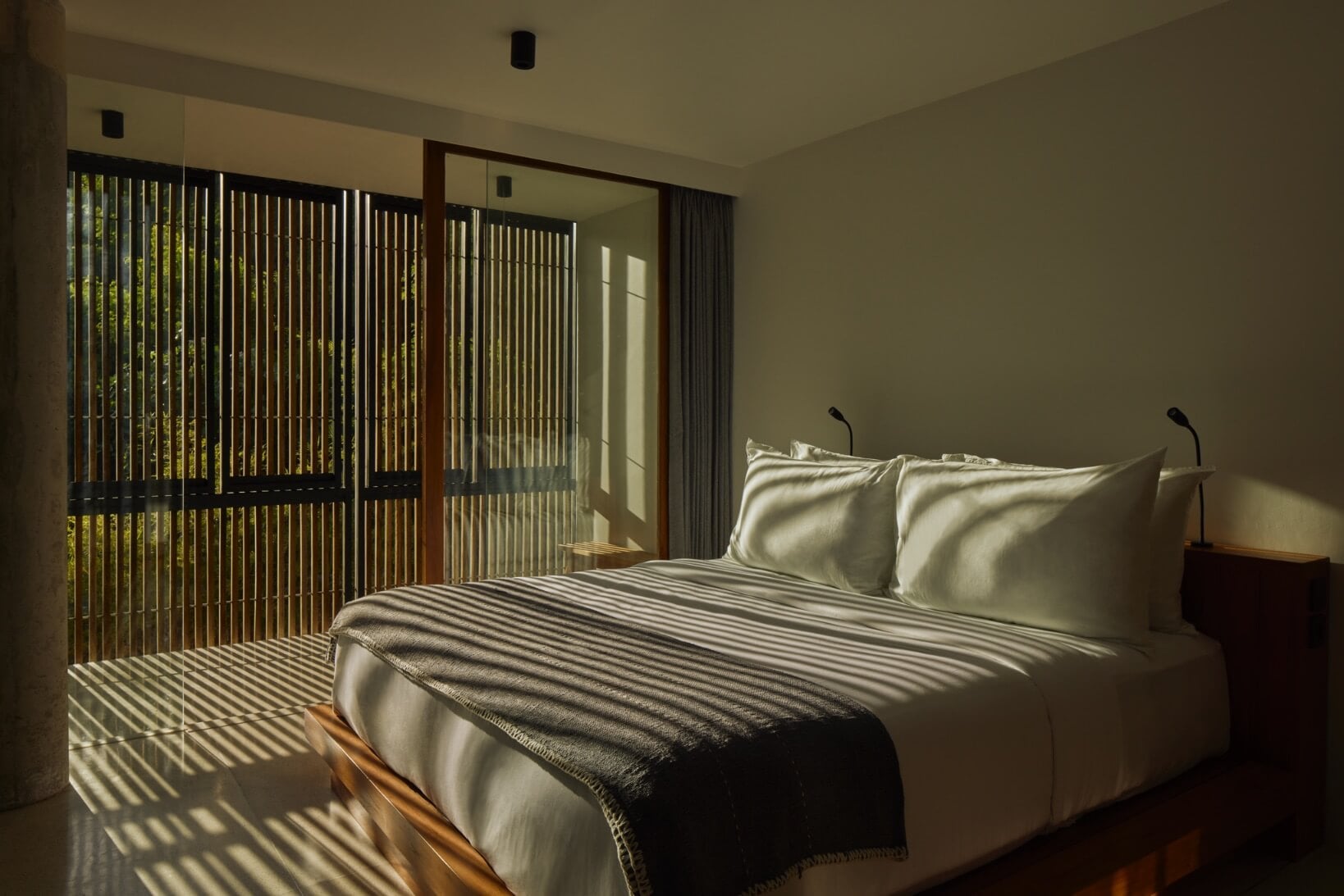 Innit Lombok, Asia — Minimalist bedroom with striped wooden screen, natural light casting linear shadows across bed with cream pillows and dark throw blanket.