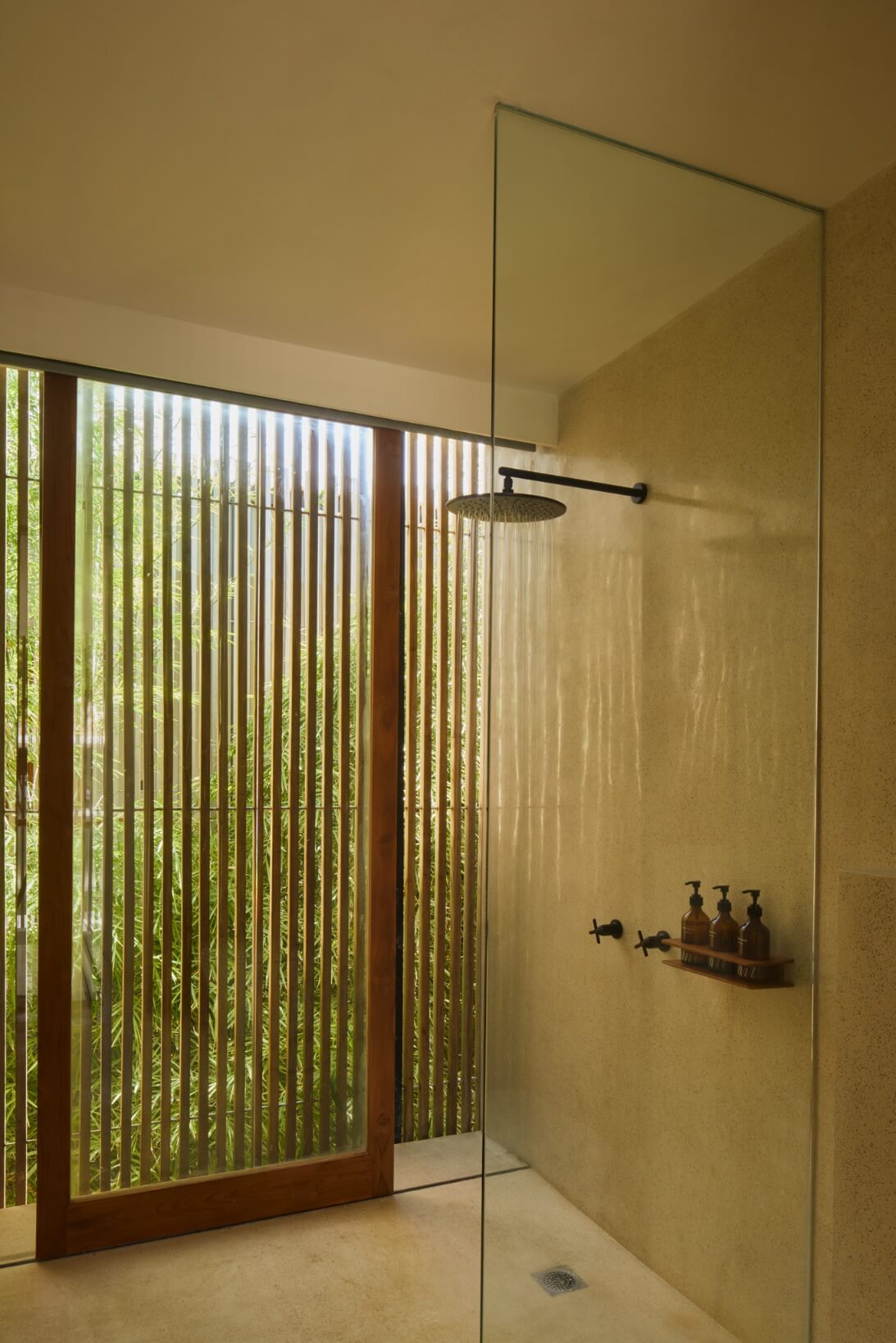 Innit Lombok, Asia — Minimalist spa shower with stone walls, rainfall showerhead, wooden louvered window framing tropical greenery, and black fixtures.