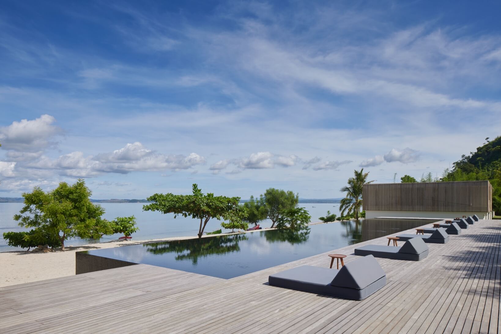 Innit Lombok, Asia — Infinity pool with dark loungers on wooden deck overlooking calm lagoon, palm trees, and distant sea under blue sky.