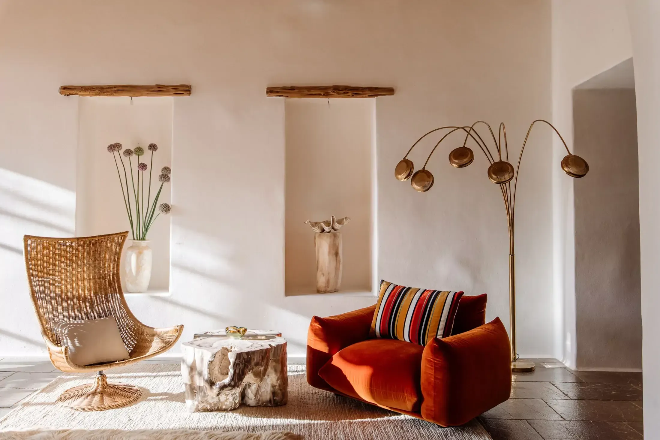 Finca La Inika, Balearic Islands, Spain — Sunlit living room with woven rattan chair, burnt-orange velvet seating, brass arc floor lamp, wooden niche shelving, and natural materials.