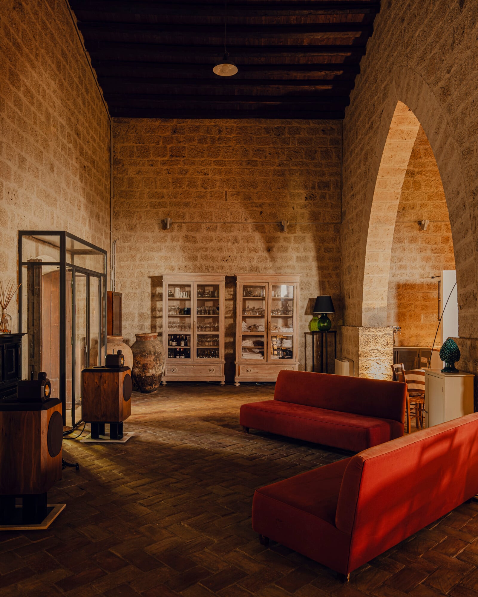 Susafa, Italy — Ochre stone gallery with vaulted arch, red modern sofas, timber ceiling, glass-front cabinetry, herringbone floor and warm ambient lighting.