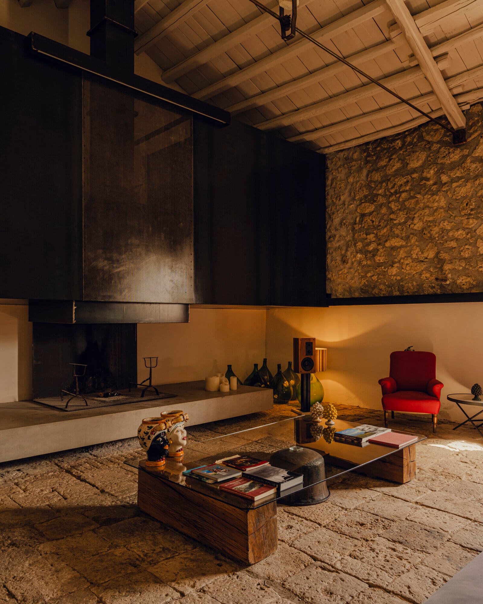 Susafa, Italy — Rustic stone interior with vaulted wood ceiling, minimalist fireplace, vintage red armchair, and ochre-toned walls bathed in warm light.