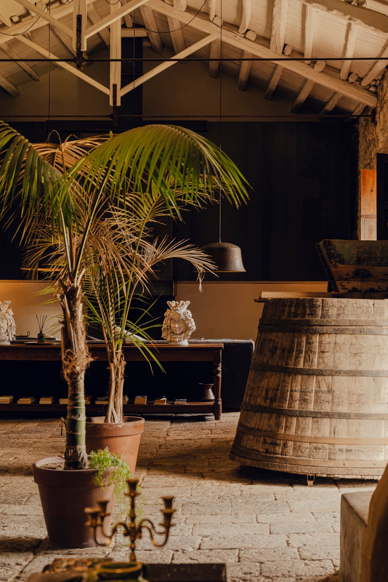 Susafa, Italy — Rustic warehouse interior with potted palm, wooden barrel, exposed beam ceiling, and ochre-walled gallery space in warm natural light.
