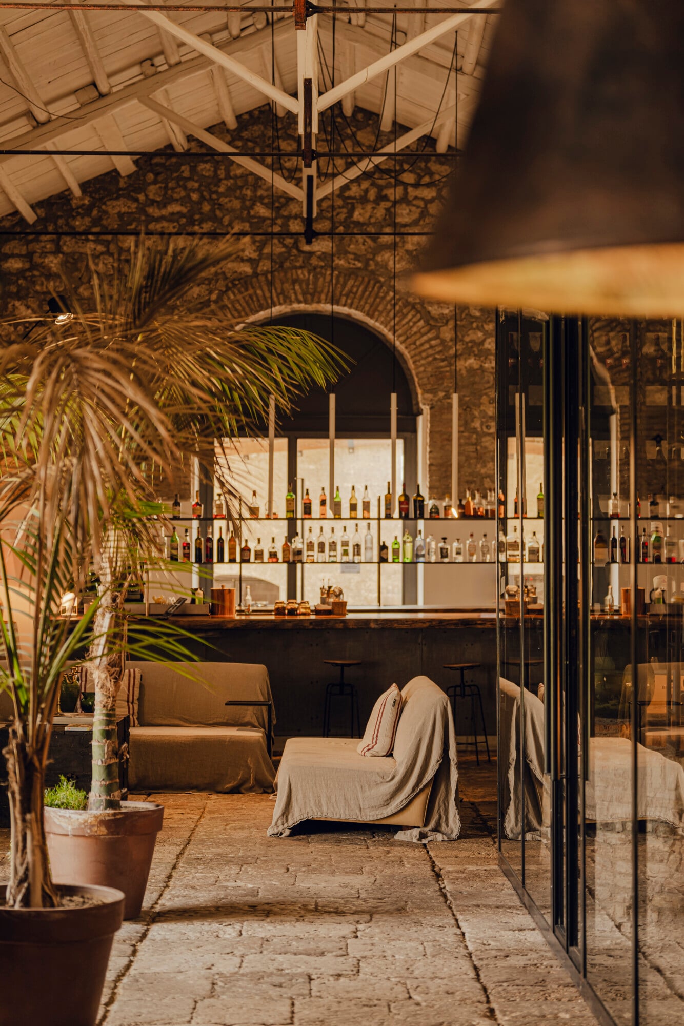 Susafa, Italy — Covered terrace with stone flooring, palm plants, cream linen seating, and backlit bar with bottle displays under timber ceiling.