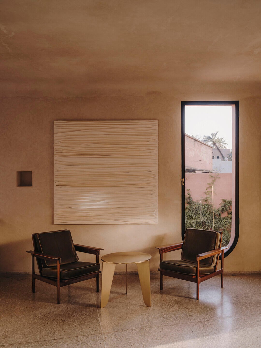 Maison Brummell Majorelle, Africa — Minimalist lounge with paired mid-century chairs, concrete side table, ochre plaster walls, wooden panel art, arched doorway opening to terracotta courtyard.
