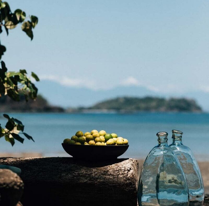 Islas Secas, Panama — Bowl of fresh limes on driftwood with glass bottles overlooking turquoise bay and forested islands under clear sky.