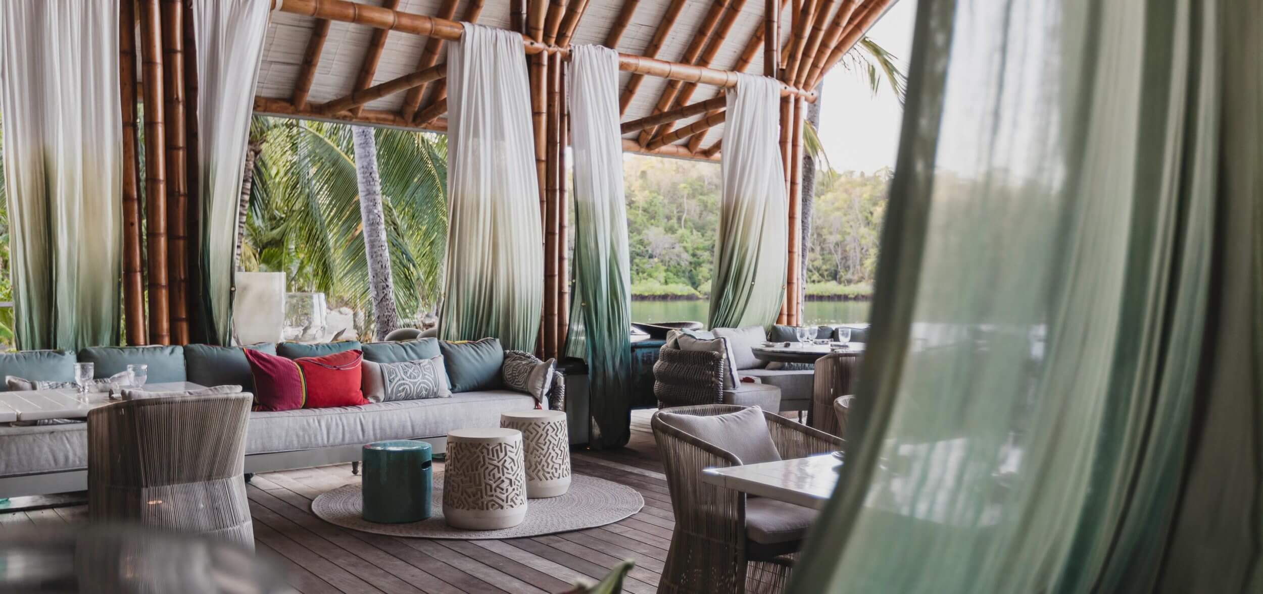Islas Secas — Waterfront pavilion with bamboo structure, flowing linen curtains, woven furniture, and ocean views through palm trees.