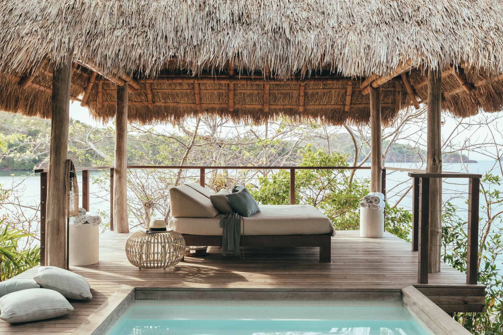 Islas Secas, Panama — Thatched-roof pavilion with wooden decking, daybed with pillows, woven poufs, and private plunge pool overlooking tropical bay and mangrove coastline.