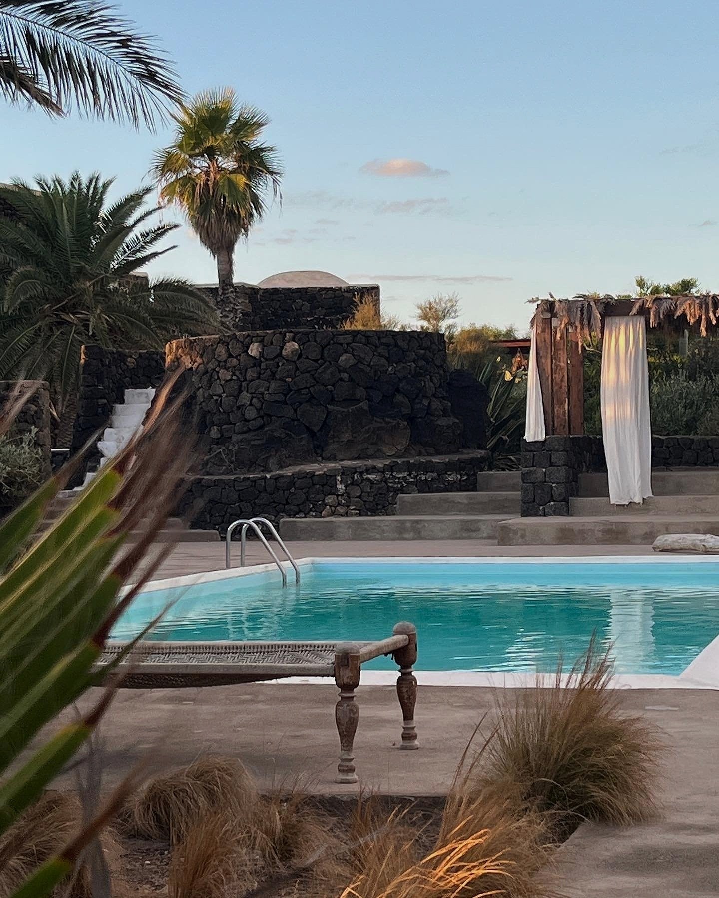 Parco Dei Sesi, Europe — Volcanic stone terraced architecture with turquoise pool, palm trees, and white draped cabanas at sunset.