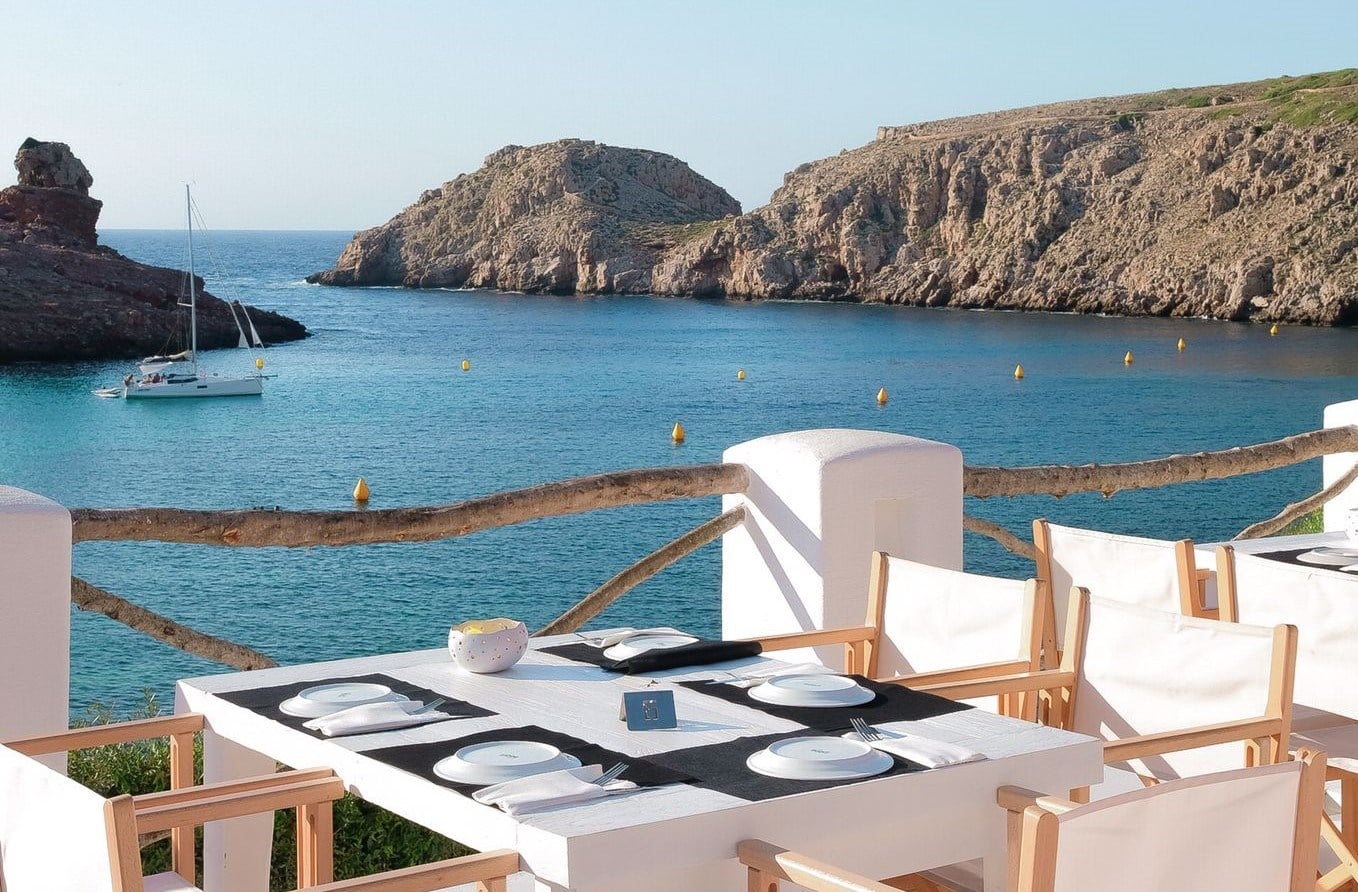 Ivette, Balearic Islands — Clifftop dining terrace with white linen table, turquoise cove, moored sailboats, and limestone rock formations.
