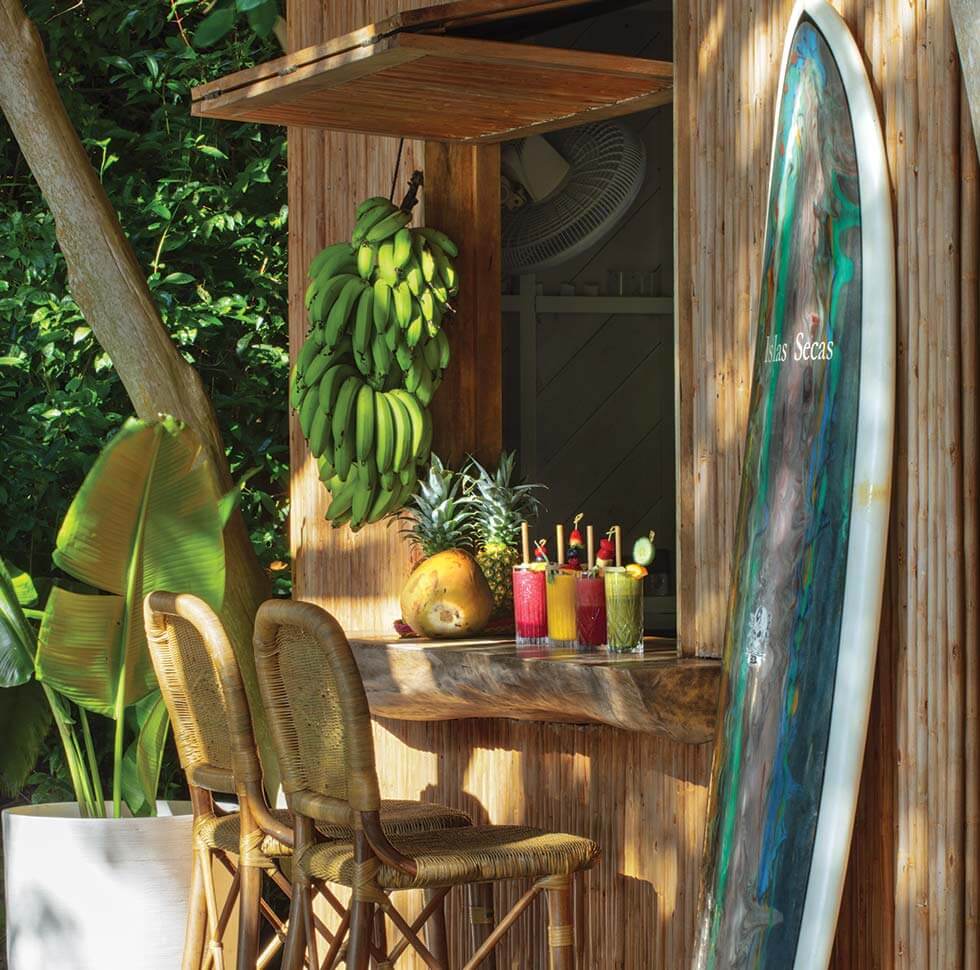 Juice Bar, All Destinations — wooden tropical bar with fresh fruit display, colorful beverages, surfboard décor, and woven bar seating in lush garden setting.