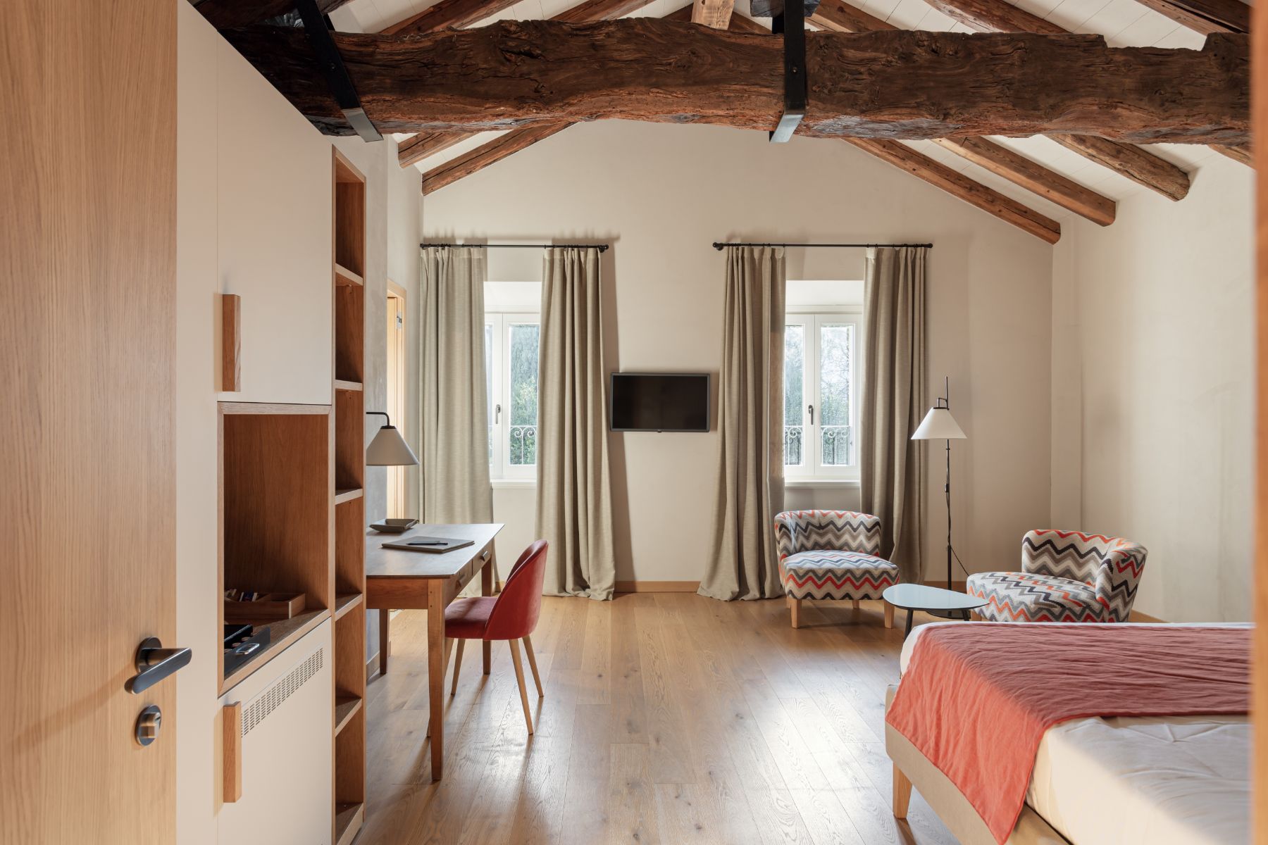 Roncolo 1888, Europe — Spacious hotel suite with exposed wooden beams, neutral linen curtains, patterned armchairs, and warm wood flooring under natural light.