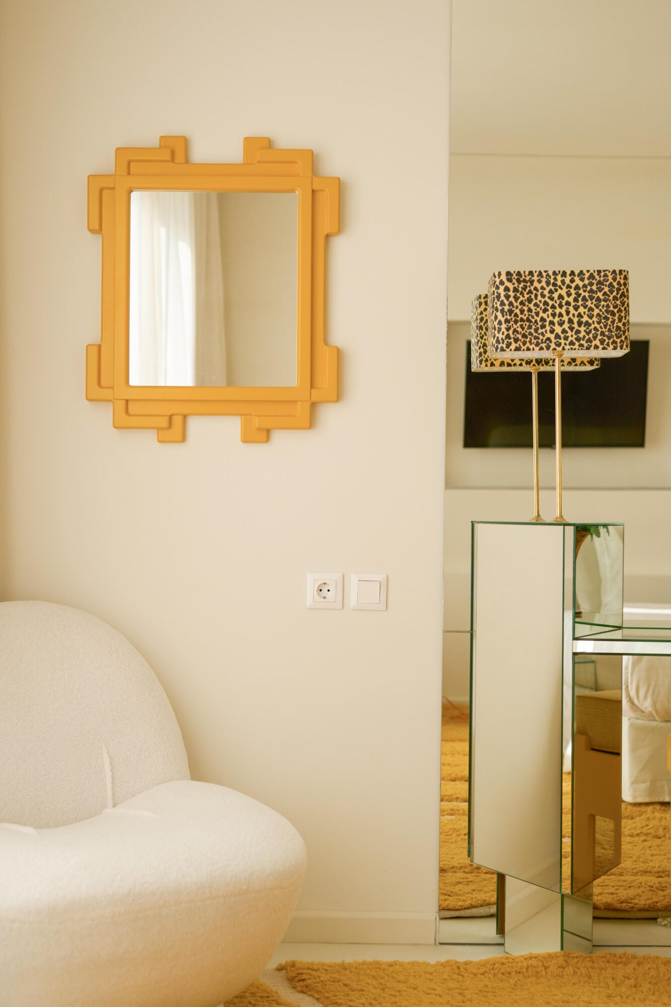 Apollo Palm, Europe — Minimalist hotel room with golden-framed geometric mirror, curved cream armchair, mirrored pedestal table with leopard-print lamp, warm natural light.