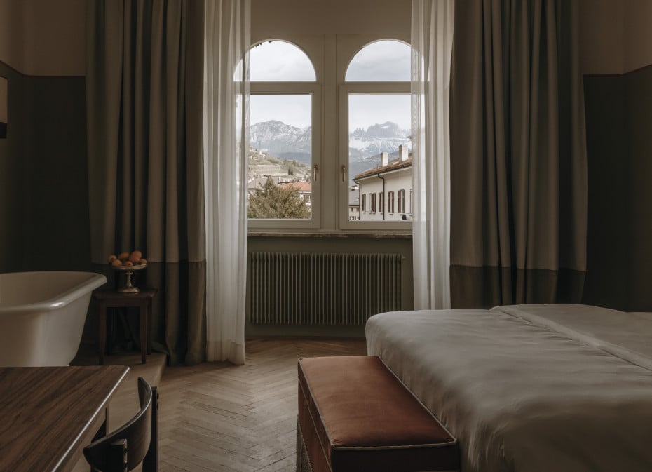 Parkhotel Mondschein, Europe — Minimalist bedroom with arched windows framing Alpine mountains, neutral linen bedding, sage radiator, layered curtains, herringbone floors, freestanding soaking tub.