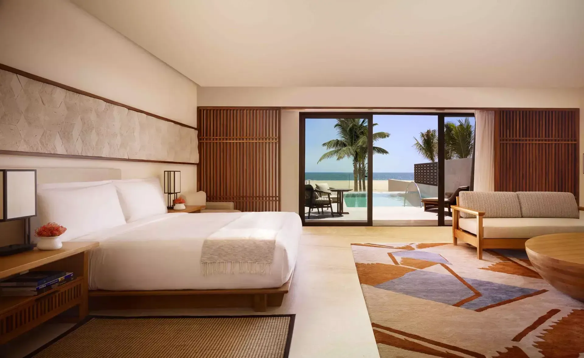 Nobu Los Cabos, Mexico — Modern beachfront suite with walnut furnishings, white bedding, geometric rugs, and floor-to-ceiling ocean views through sliding glass doors.