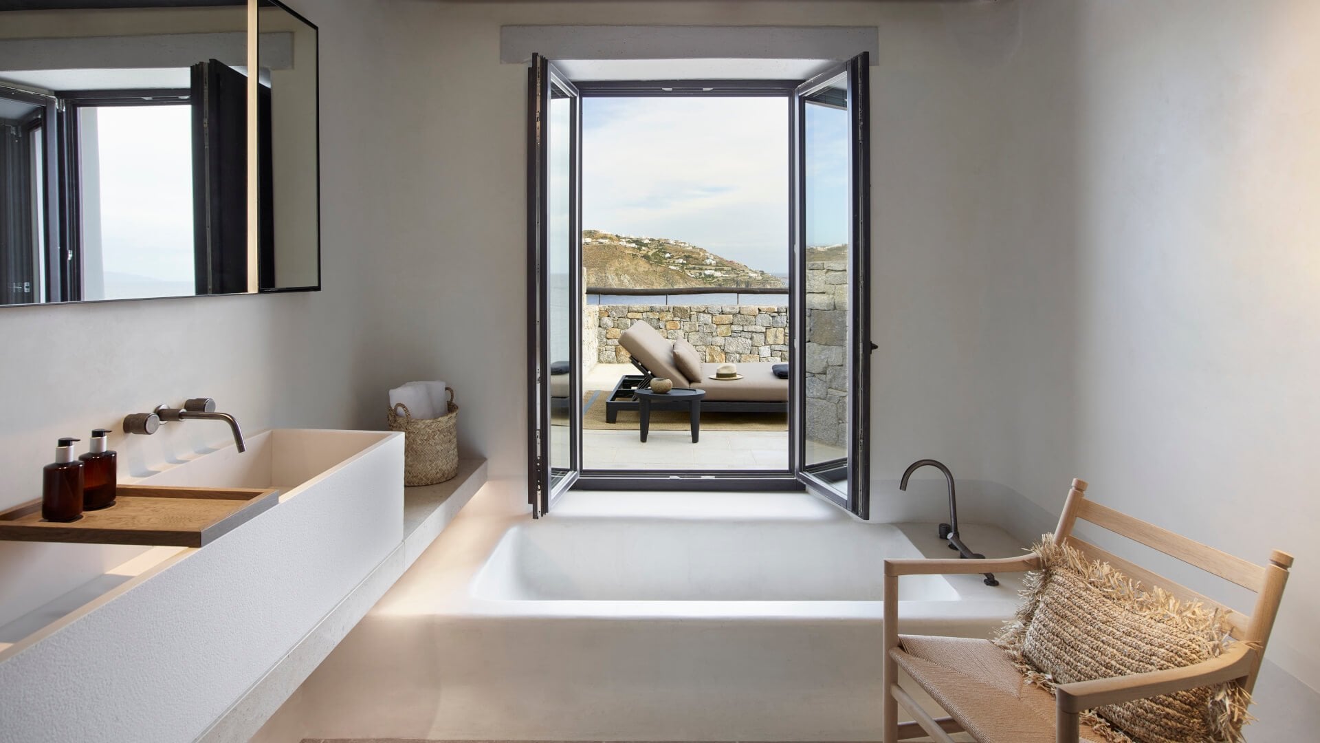 Kalesma, Europe — Minimalist bathroom with stone terrace view, sculptural white bathtub, natural wood accents, and hillside landscape through open doors.