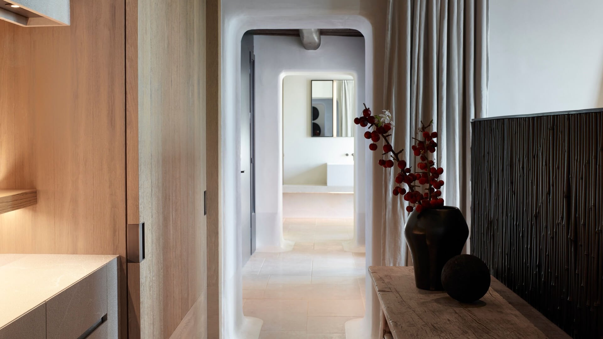 Kalesma, Europe — Minimalist hallway with white archway, warm wood cabinetry, dark ceramic vessels with red branches, natural light, luxury hospitality design.