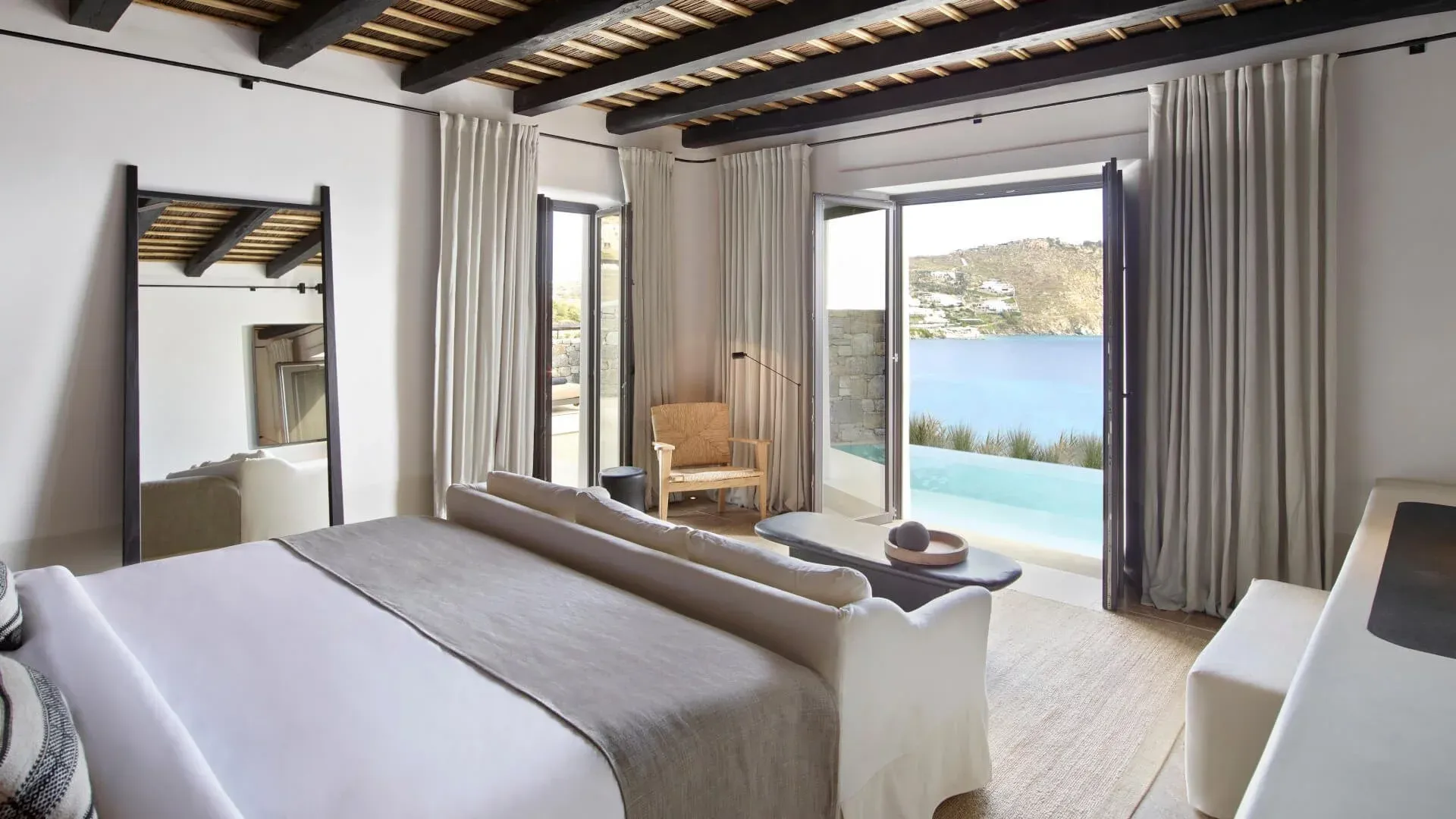 Kalesma, Europe — Minimalist bedroom suite with coastal views, wooden ceiling beams, cream linen furnishings, and infinity pool overlooking Mediterranean landscape.