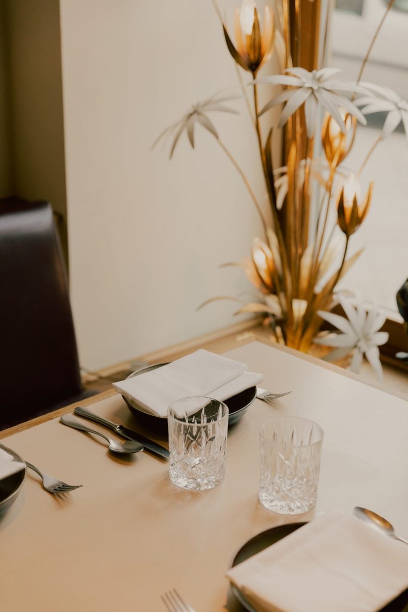 Kin Dee, Europe — Elegant table setting with crystal glassware, white linens, dark cutlery, and dried gold floral arrangement on neutral backdrop.