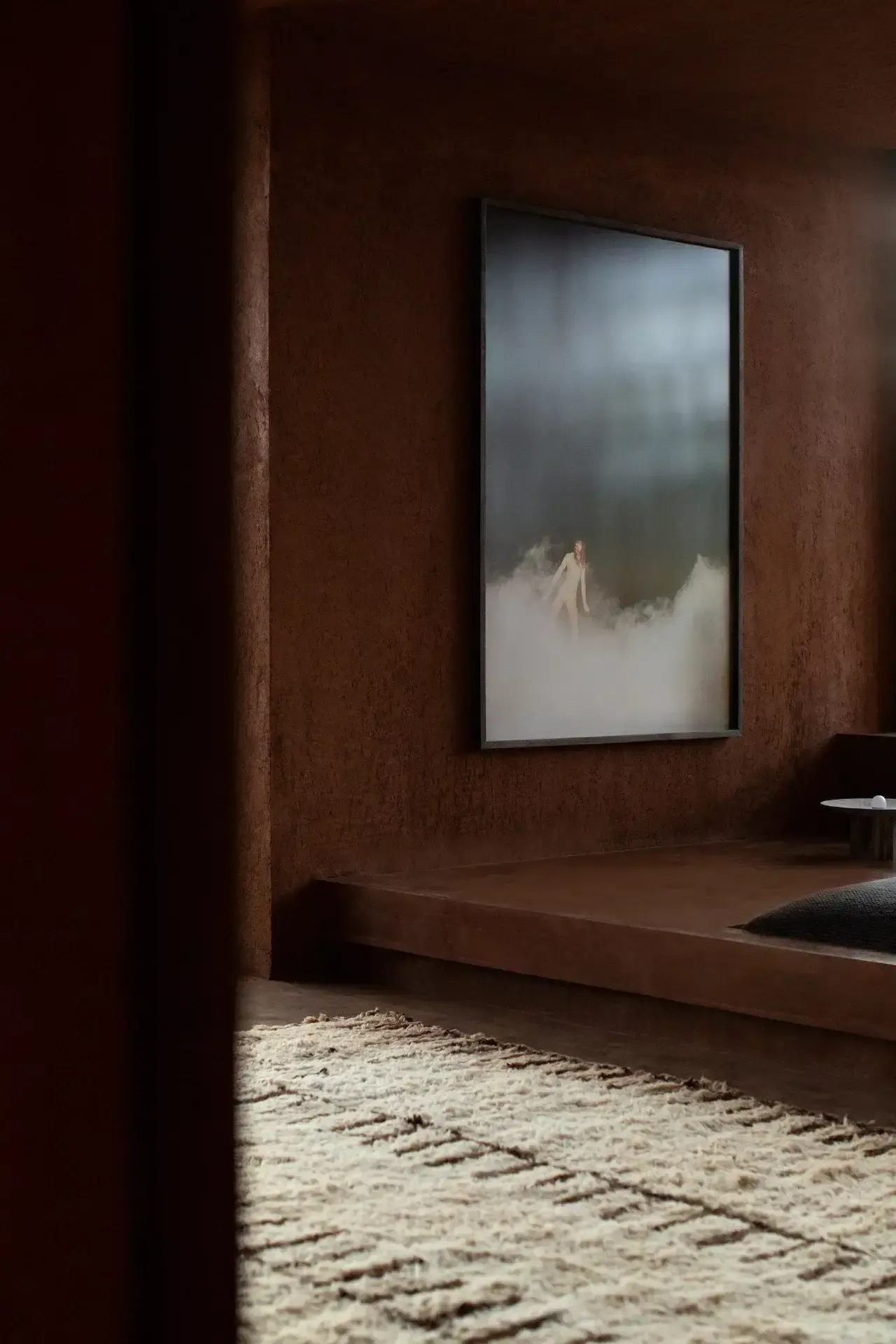 Further Hotel, Asia — Minimalist room with terracotta walls, concrete platform bed, and framed mountain landscape artwork with soft natural light casting shadows across textured flooring.