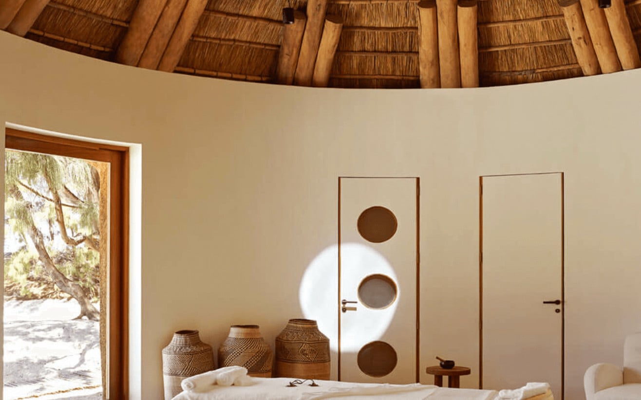 Natural Wellness Centre, Africa — Circular thatched-roof spa room with woven baskets, minimalist white door with circular apertures, wooden beam ceiling, and soft natural light from large opening.