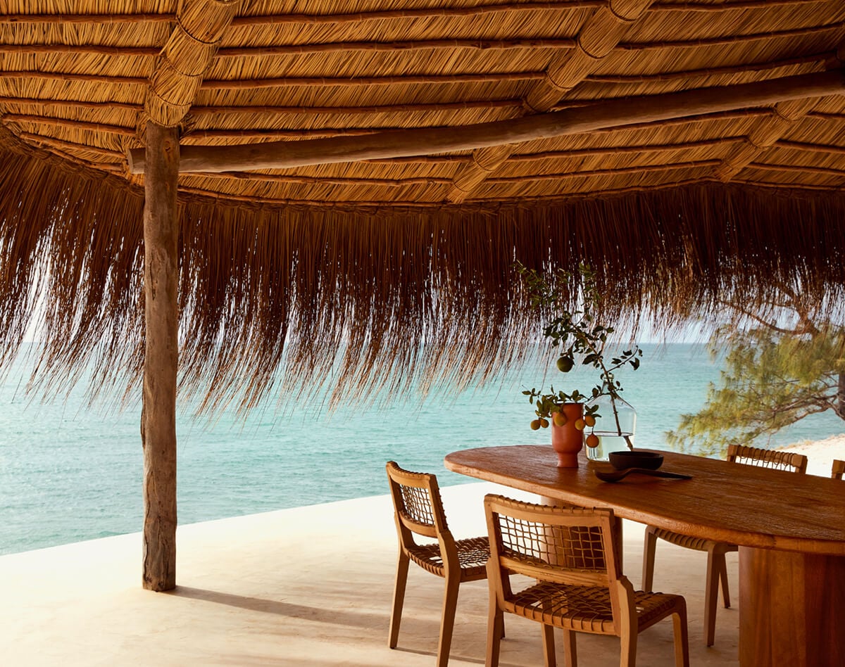 Kisawa Sanctuary, Africa — Thatched-roof pavilion with wooden table and woven chairs overlooking turquoise ocean and white sand beach.