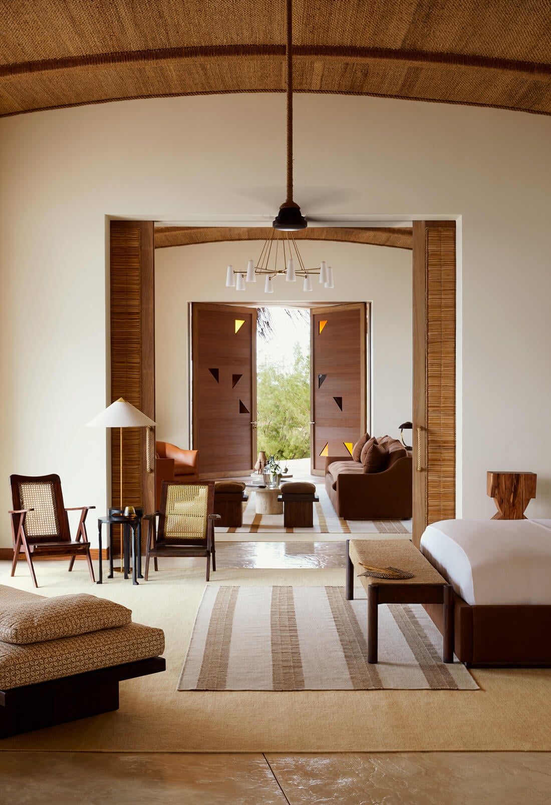 Kisawa Sanctuary, Africa — Minimalist bedroom suite with arched timber ceiling, wooden louvered doors opening to living pavilion, striped natural fiber rug, mid-century furniture, and diffused tropical light.