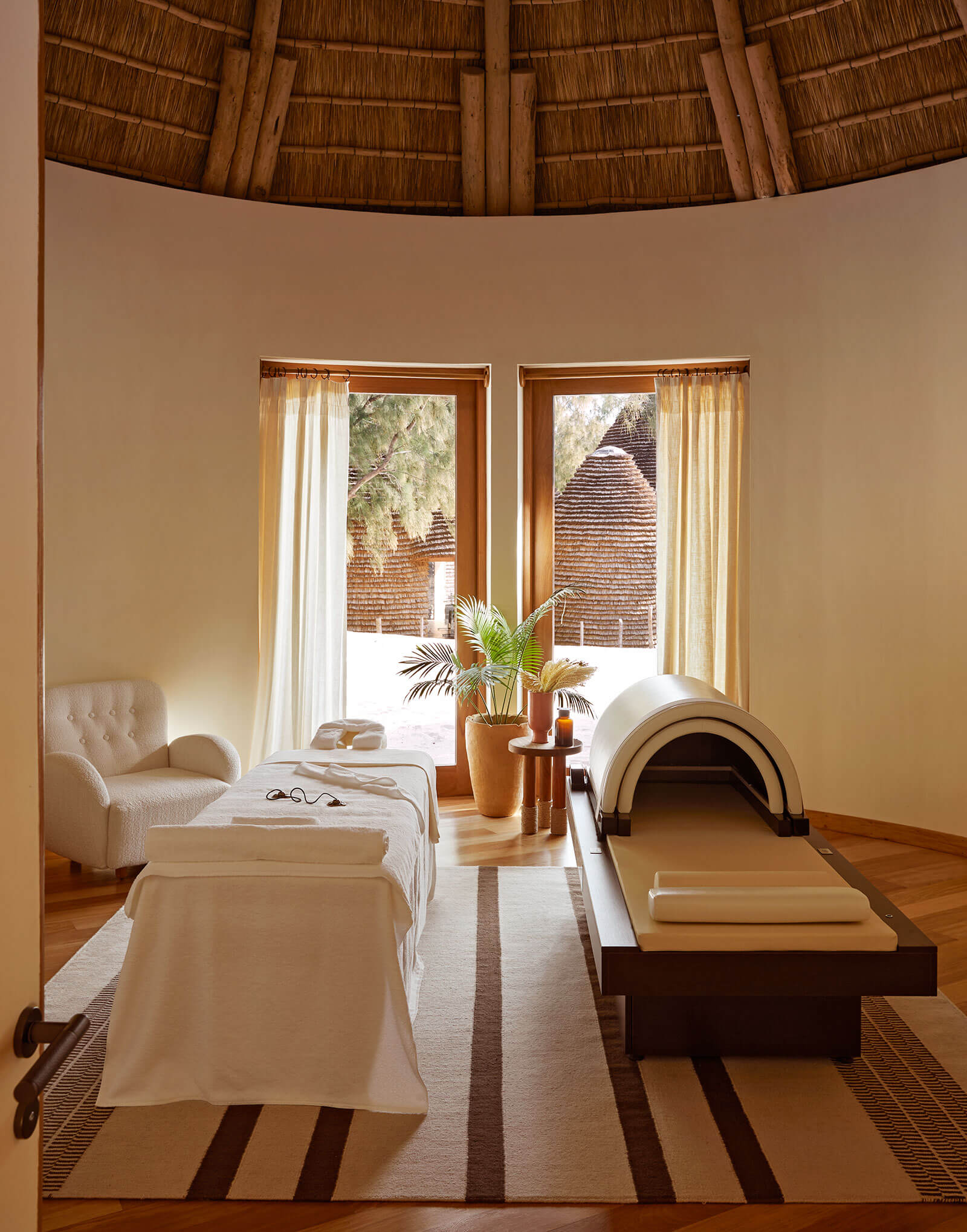 Iyashi Dome, Africa — Spa treatment room with thatched dome ceiling, massage tables, cream armchair, potted plant, and brick structures visible through floor-to-ceiling doors.