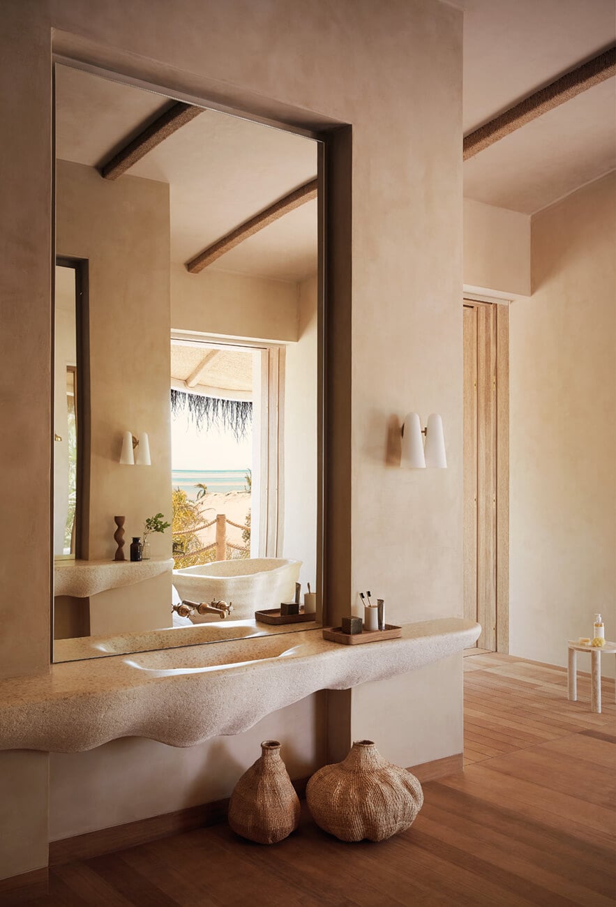 Kisawa Sanctuary, Africa — Minimalist bathroom with curved limestone sink, large mirror, exposed wooden beams, and ocean view through open terrace doors.