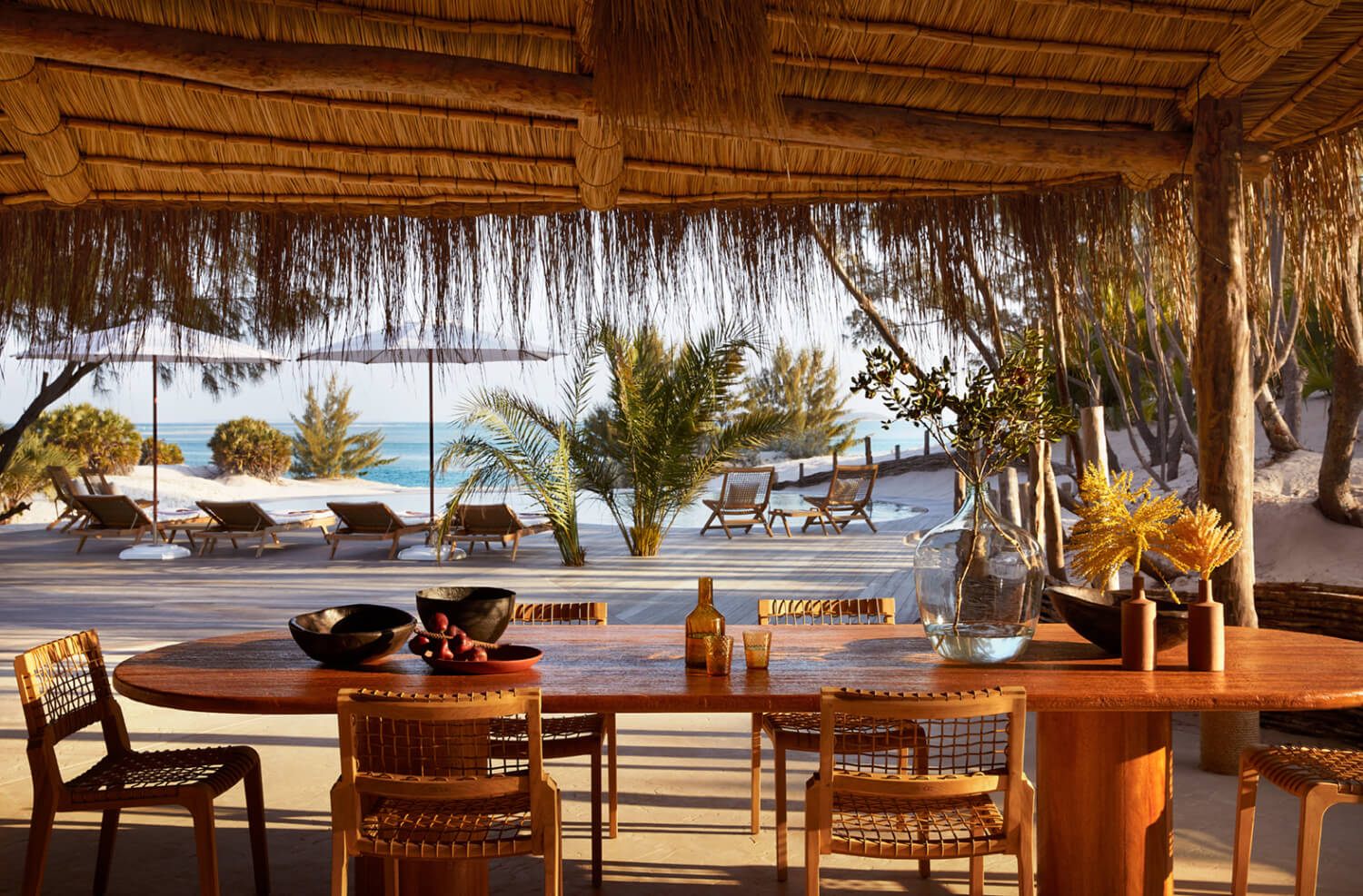 Kisawa Sanctuary, Africa — Thatched pavilion with wooden dining table overlooking palm-lined beach and turquoise ocean, natural light filtering through woven roof.
