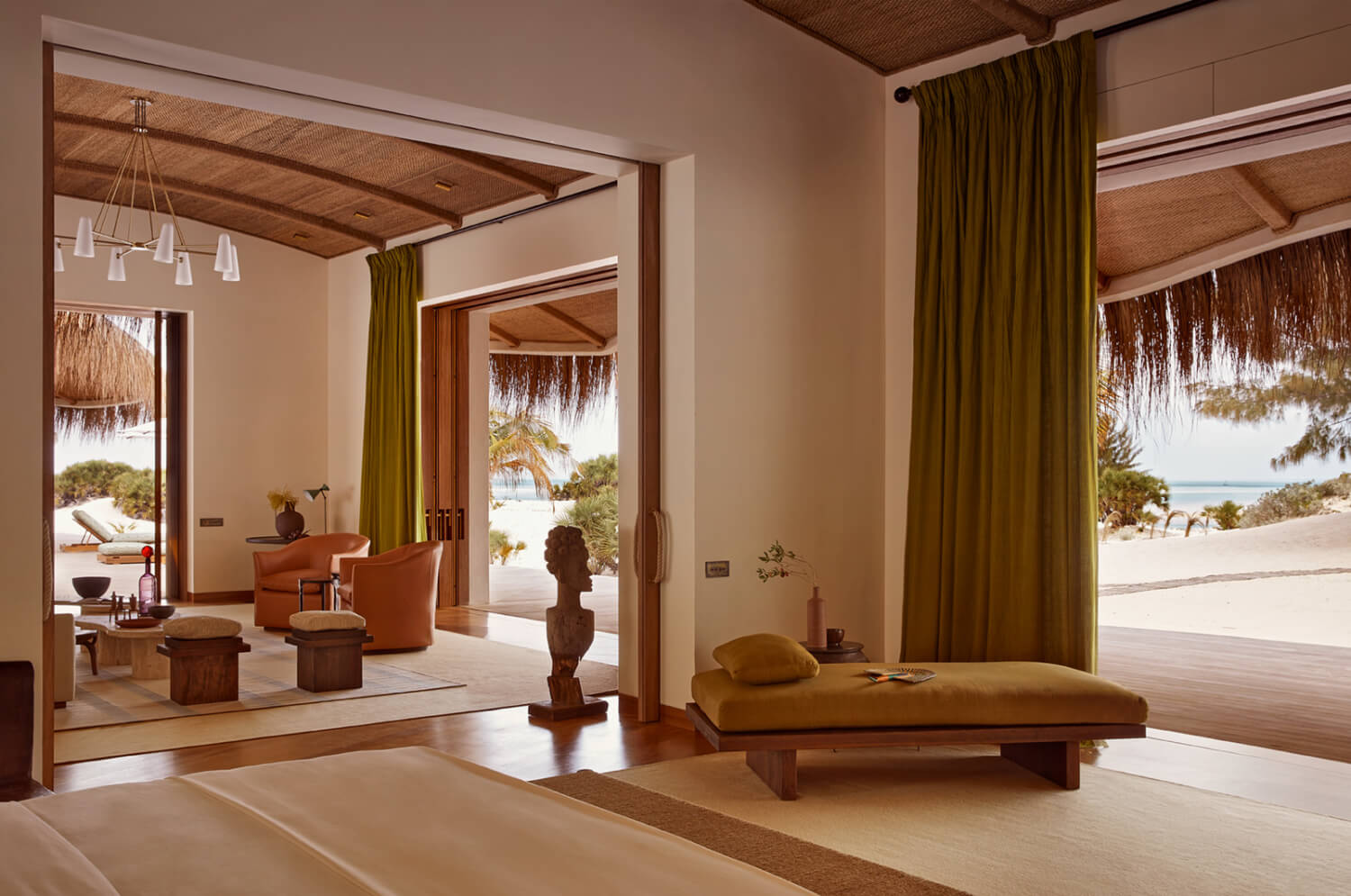 Kisawa Sanctuary, Africa — Minimalist bedroom with warm earth tones, wooden platform bed, olive curtains, open terraces overlooking white sand beach and ocean.
