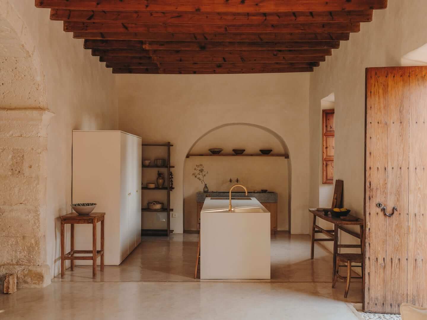 Can Lluïssó, Balearic Islands, Spain — Minimalist kitchen with arched niche, white island, exposed wooden beams, terracotta floors, and rustic wooden doors.