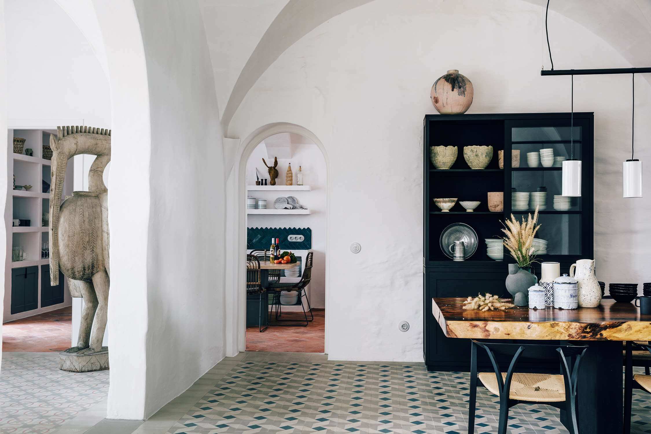 Es Bec d’Aguila — Es Bec d'Aguila, Balearic Islands, Spain — Whitewashed arched interior with black cabinetry, terracotta tile floors, and sculptural stone figure by entrance.