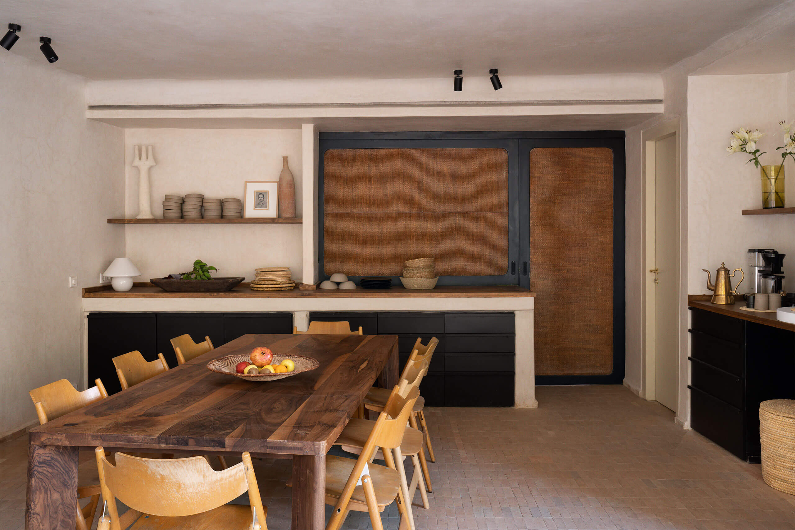 Maison Brummell Majorelle, Africa — Minimalist dining room with walnut table, woven rattan cabinet doors, cream plaster walls, wooden chairs, and curated shelf styling.