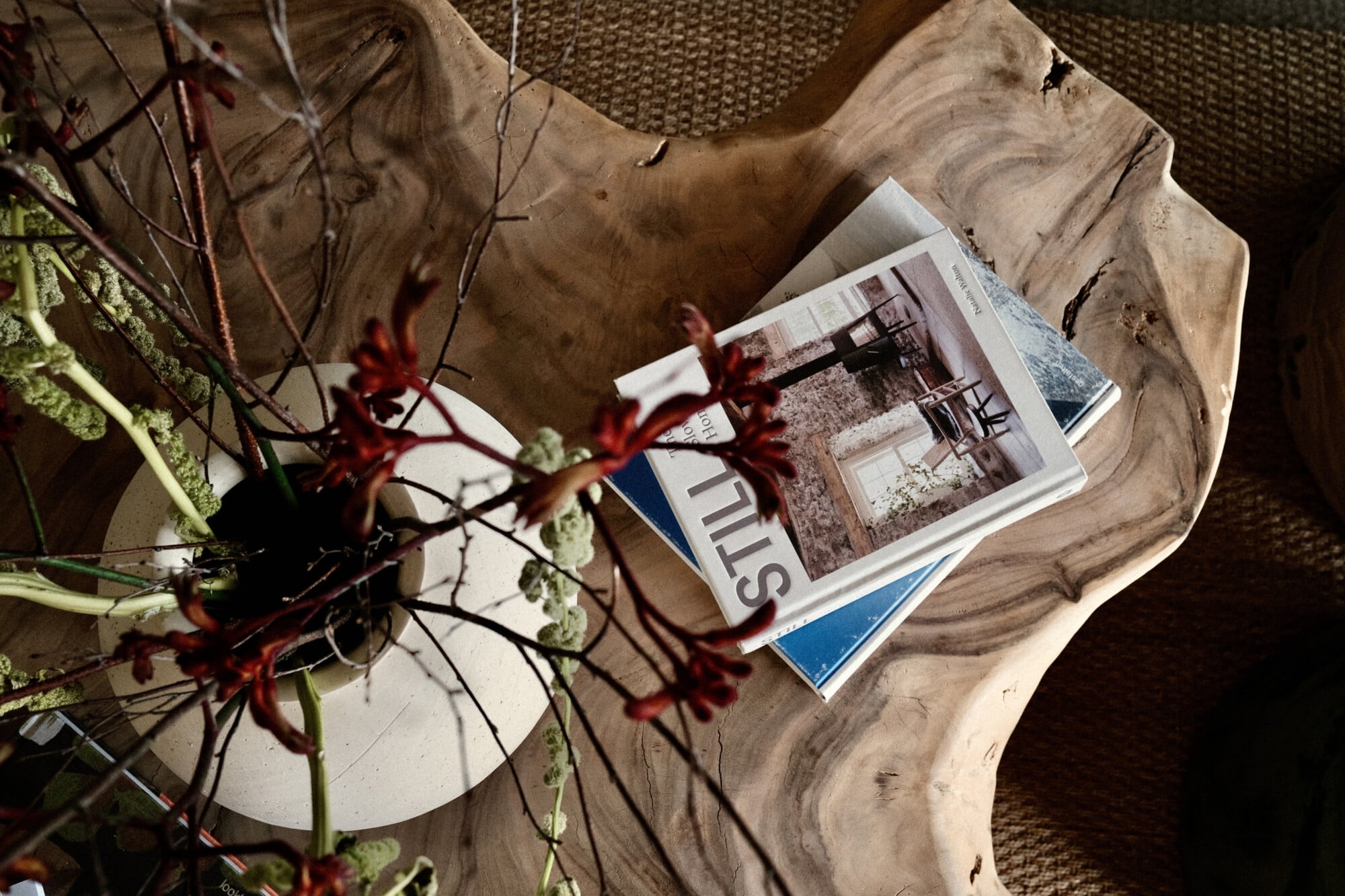 Vista Verde, Balearic Islands, Spain — Wooden side table with design magazines and dark red flowers, natural light and textured fabric chair detail.