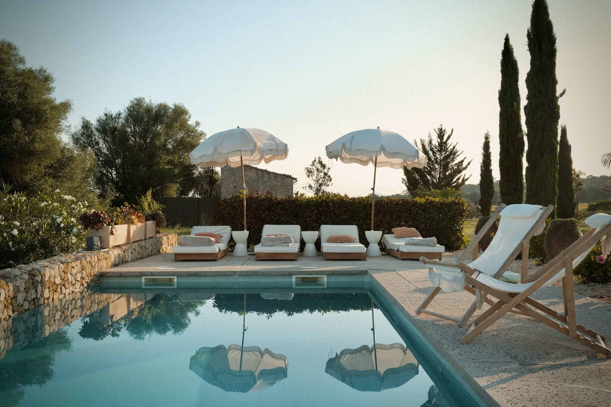 Vista Verde, Balearic Islands — Turquoise pool with stone terrace, linen sunbeds under parasols, cypress trees, afternoon light reflecting on water.