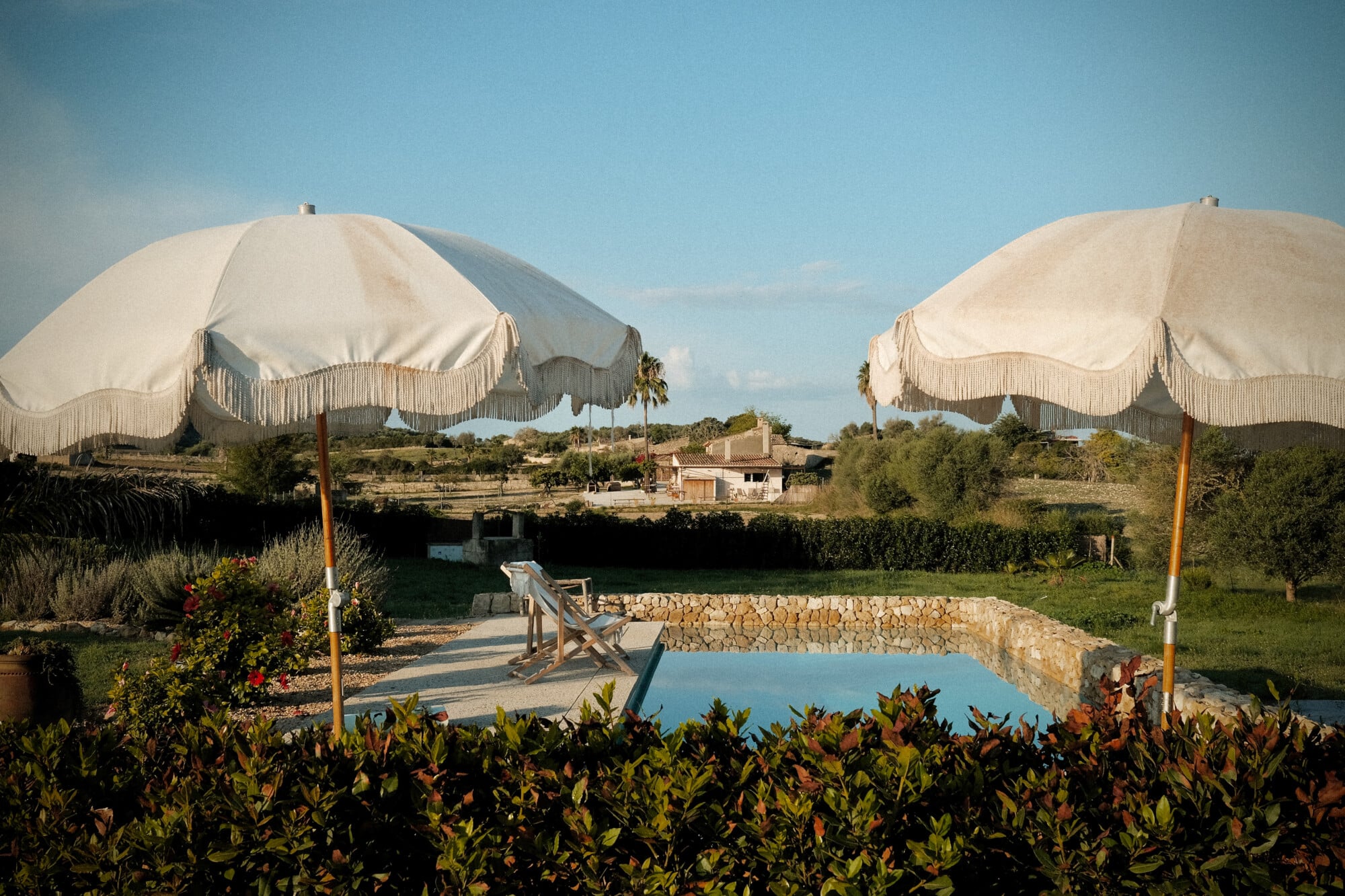 Vista Verde, Balearic Islands — Cream umbrellas with fringe shade lounge chairs beside turquoise pool, stone terrace, Mediterranean landscape beyond.