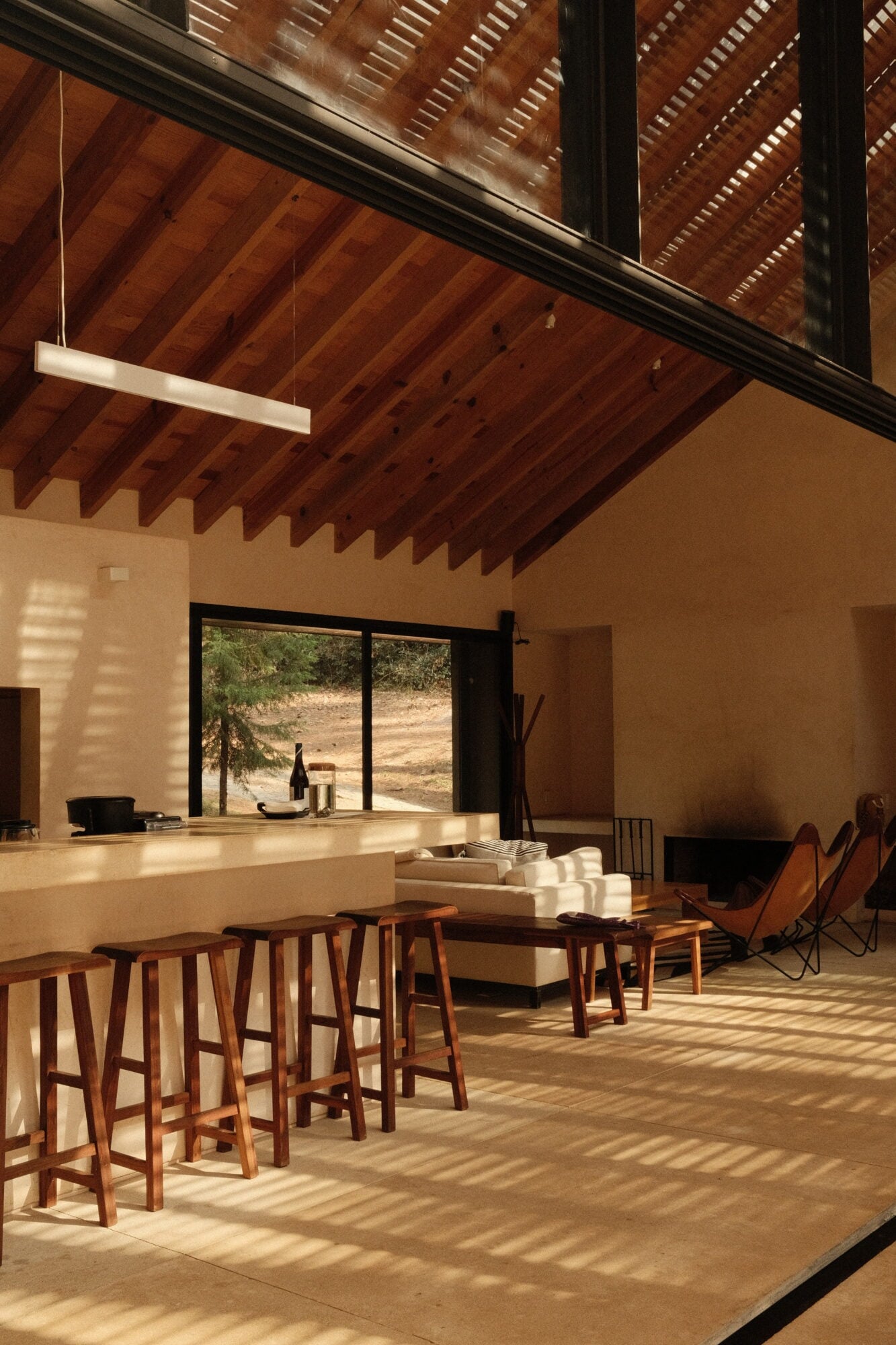 Casa Zarzales, Mexico — Minimalist open-plan living space with exposed timber beams, concrete kitchen island, wooden bar stools, and floor-to-ceiling windows overlooking hillside landscape in warm afternoon light.