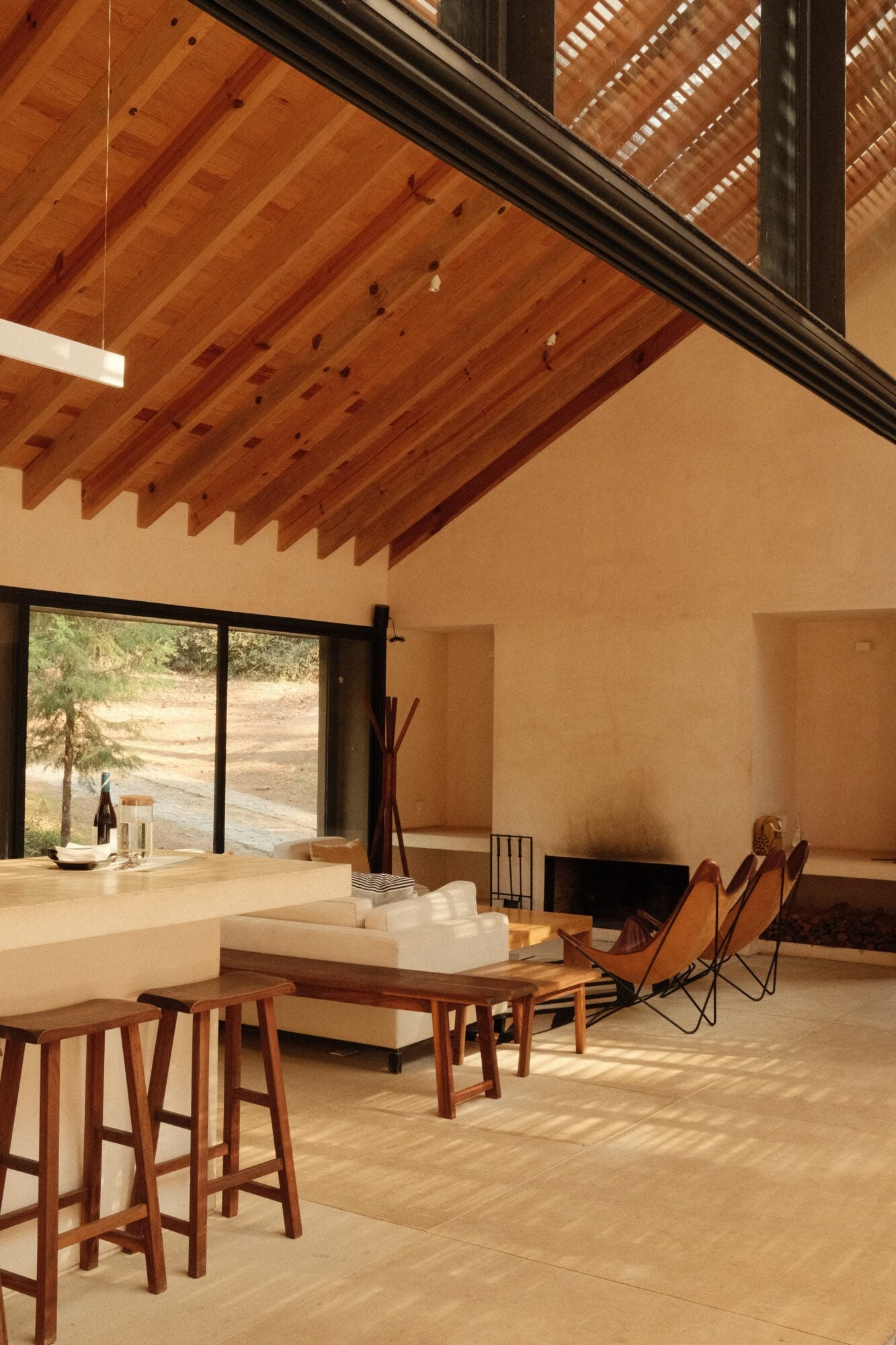 Casa Zarzales, Mexico — Open-plan living room with exposed wooden ceiling beams, terracotta walls, leather butterfly chairs, and floor-to-ceiling windows overlooking landscape.