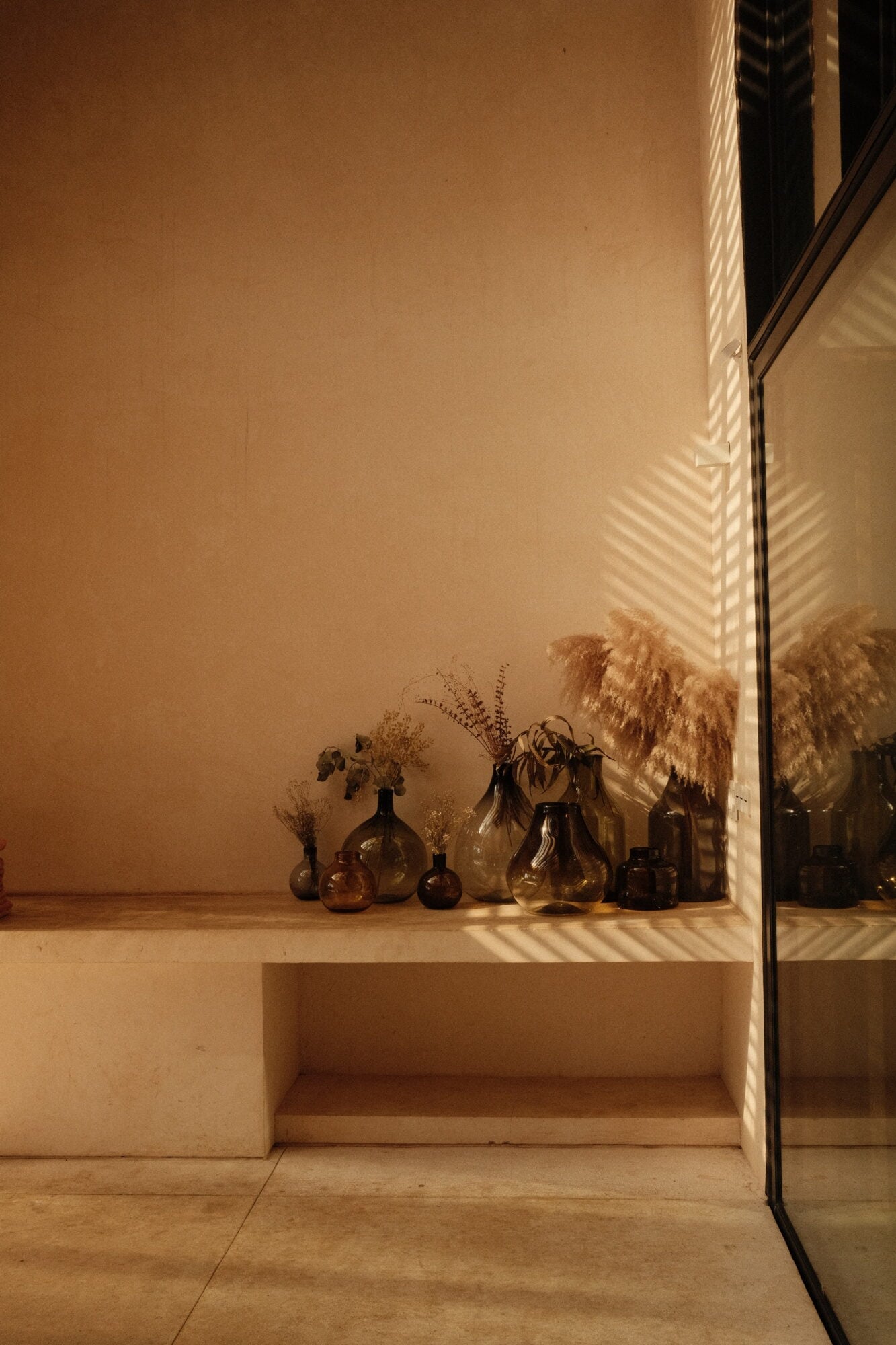 Casa Zarzales, Mexico — Minimalist interior with amber glass vessels on floating shelf, dappled sunlight casting geometric shadows on warm stone walls.
