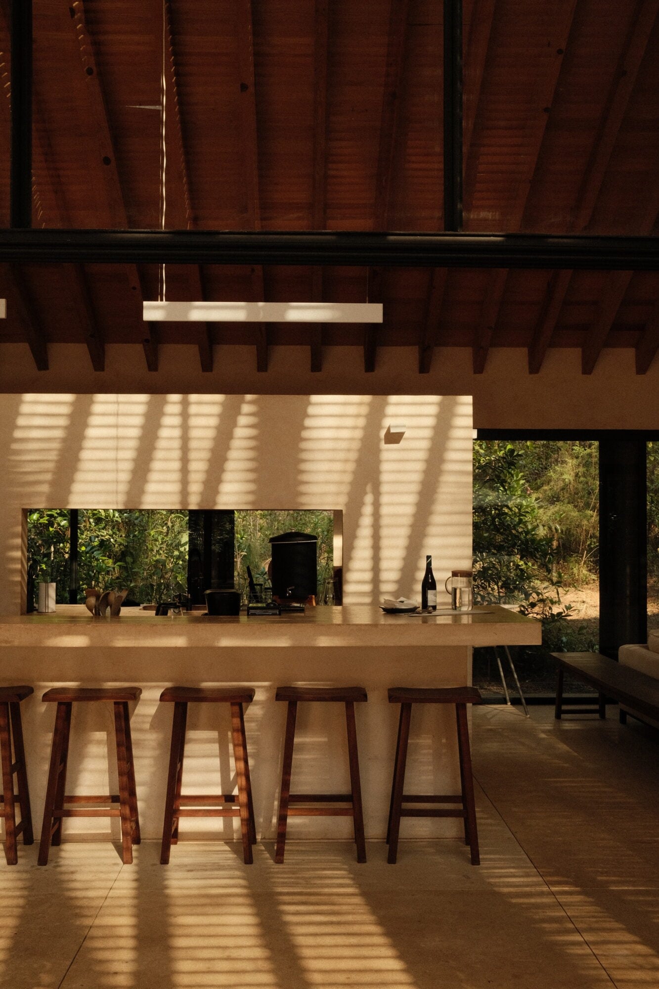Casa Zarzales, Mexico — Minimalist kitchen bar with wooden stools, concrete countertop, and clerestory windows casting geometric shadows across open-air pavilion overlooking forest.